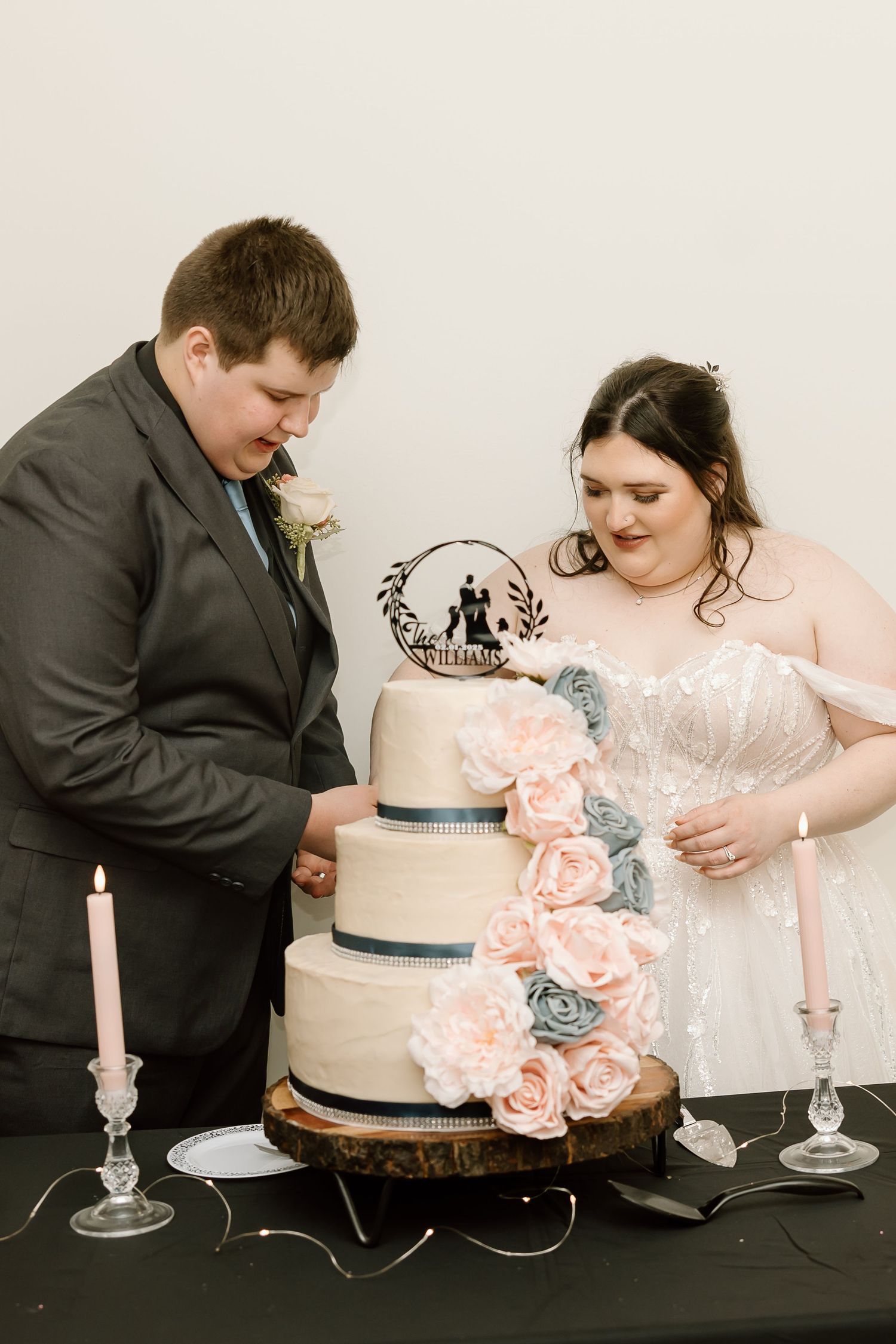 A couple cuts into their elegant white wedding cake decorated with pink and blue flowers and ribbon.