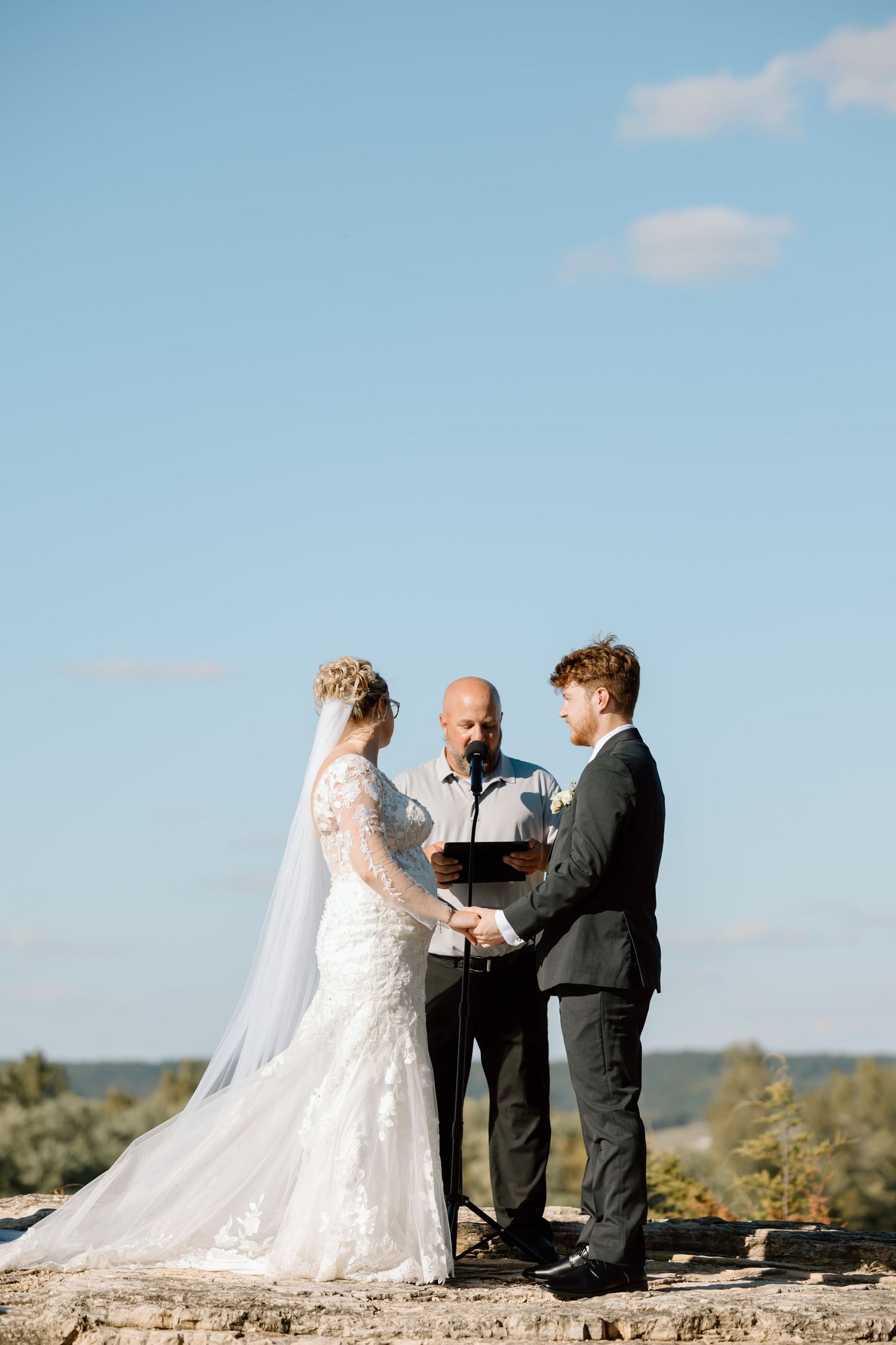 A wedding ceremony takes place on a scenic cliff overlooking a natural landscape on a sunny day.