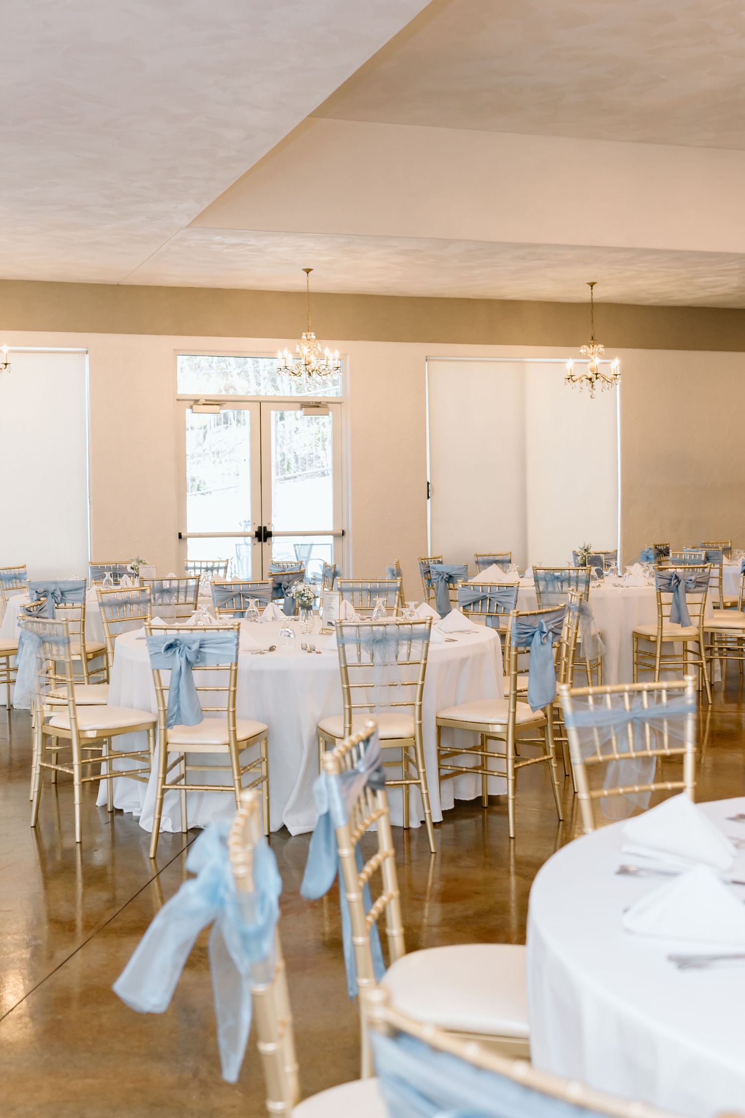 Elegant wedding reception venue with white tablecloths, gold chiavari chairs and light blue sashes in a bright banquet room.