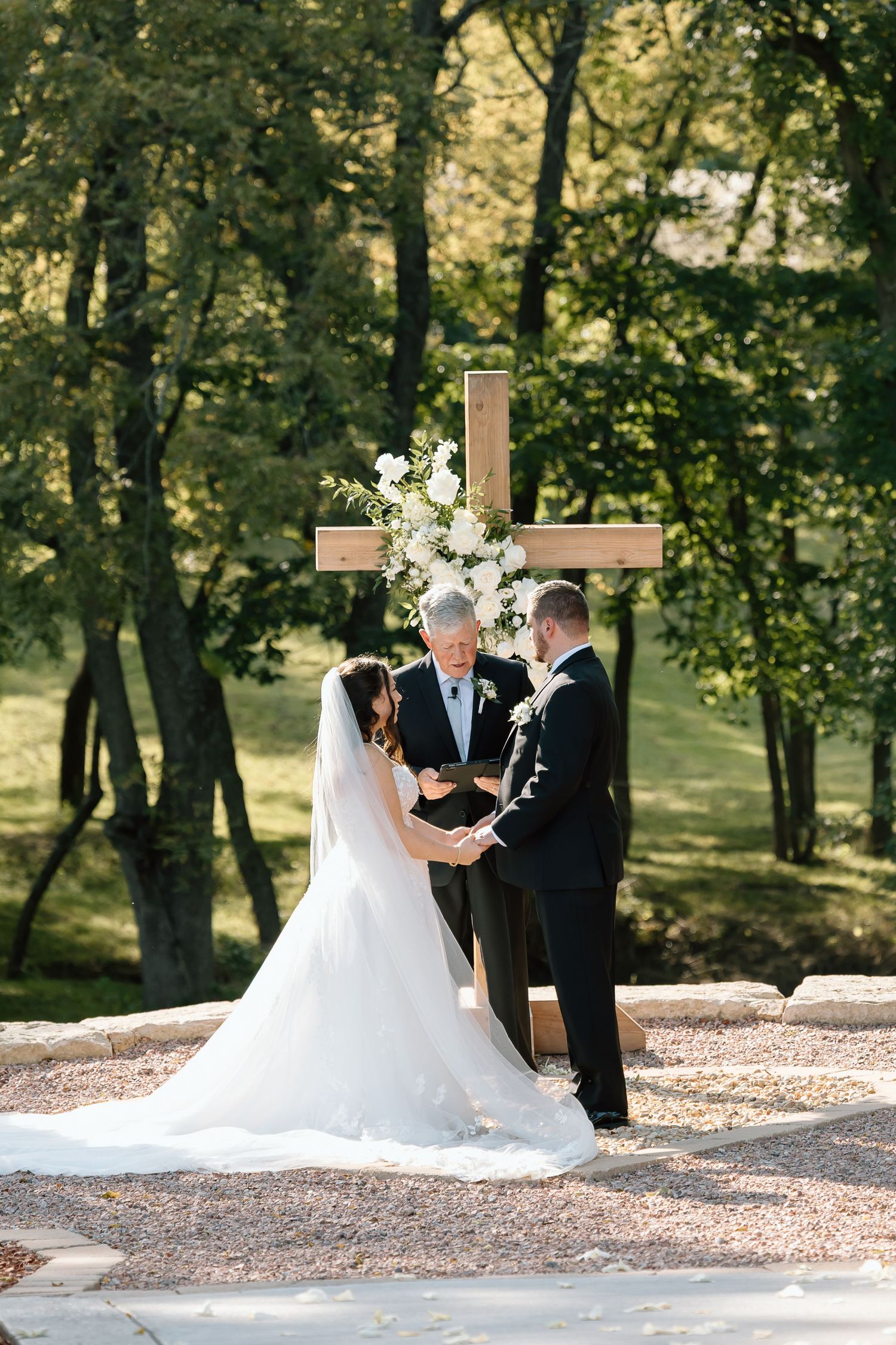 A wedding ceremony takes place outdoors in a wooded setting with a wooden cross and white floral arrangements.