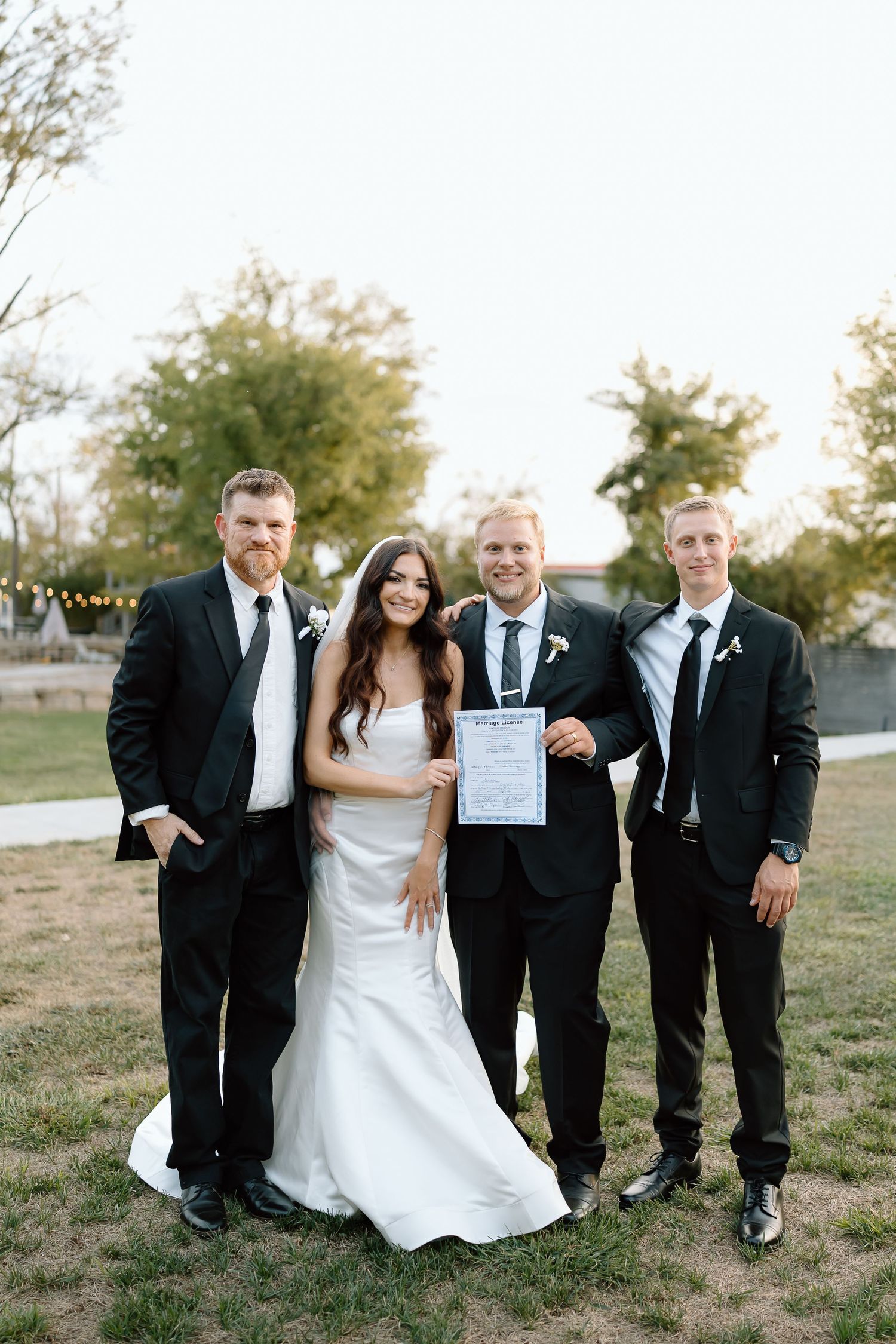 A group of people in formal attire pose together outdoors at a wedding celebration while holding a document.