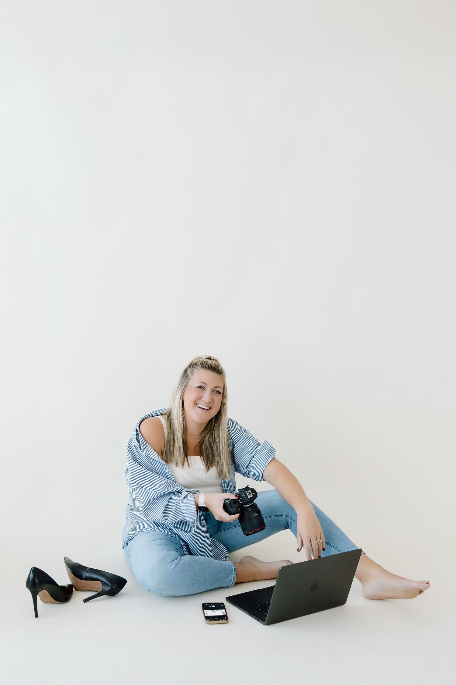 Professional photographer in light blue outfit lying on floor with camera and laptop in minimal white studio setting.
