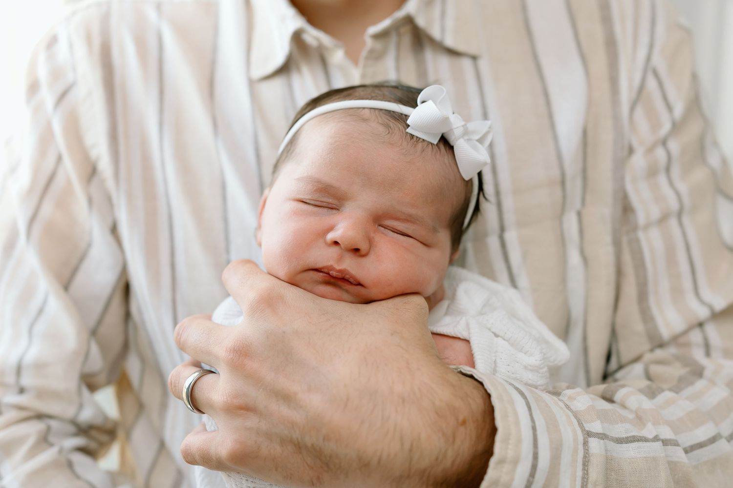 A peaceful sleeping newborn baby wearing a white bow headband is cradled in gentle arms.