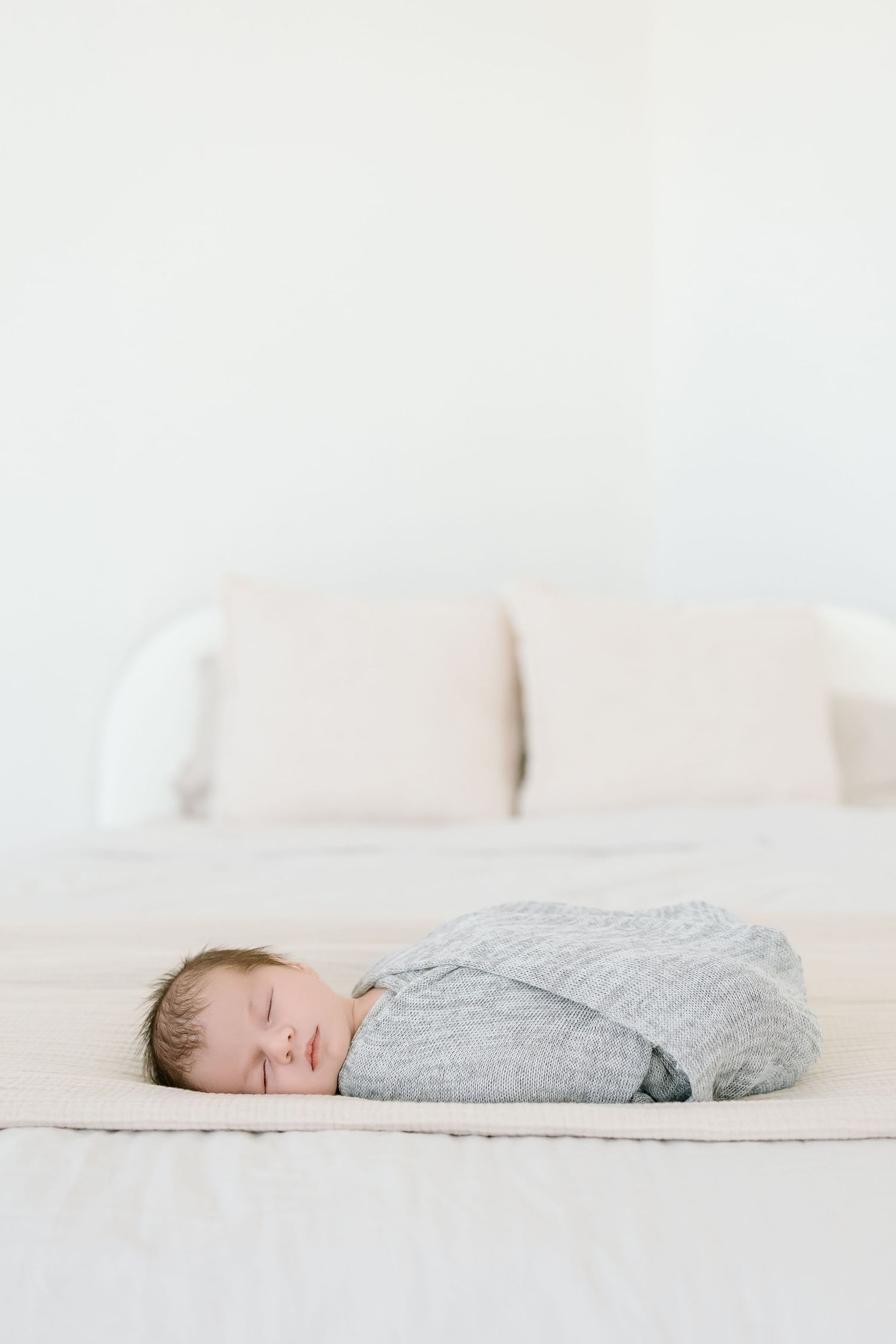 A baby wearing a gray sweater sleeps peacefully on white bedding in a minimalist setting.