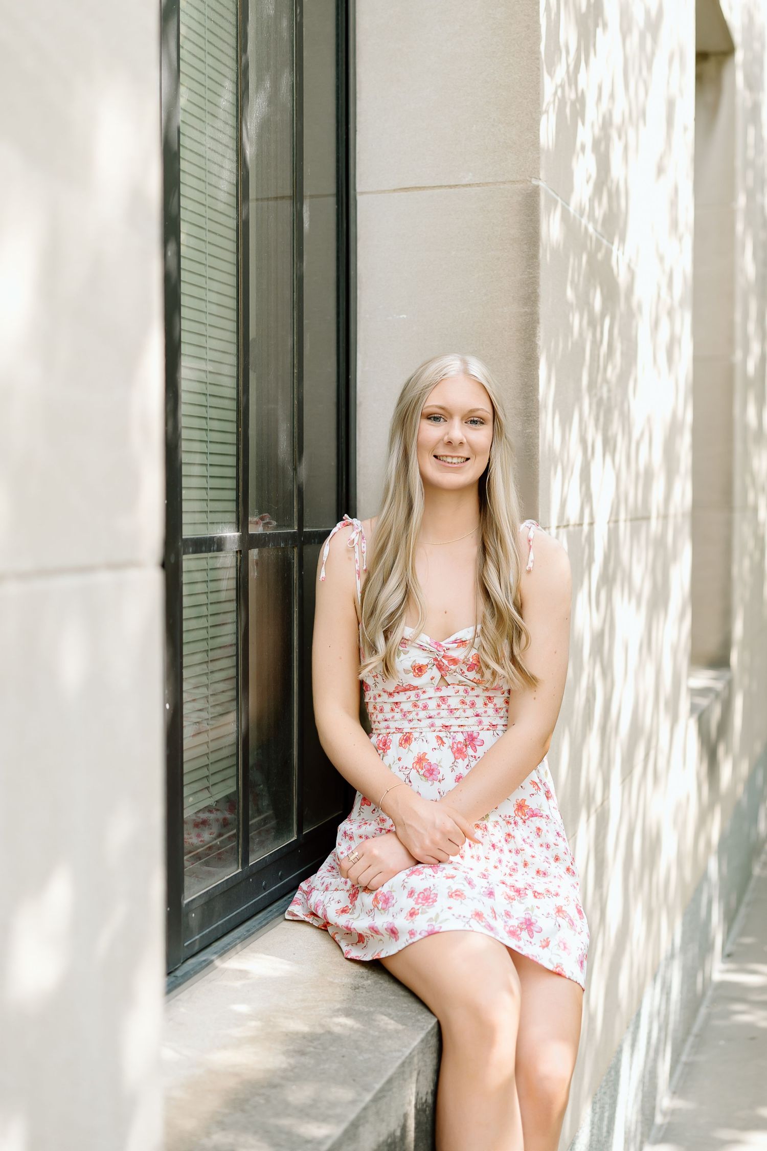 A person in a pink and white floral dress poses against a white wall with green shuttered windows.