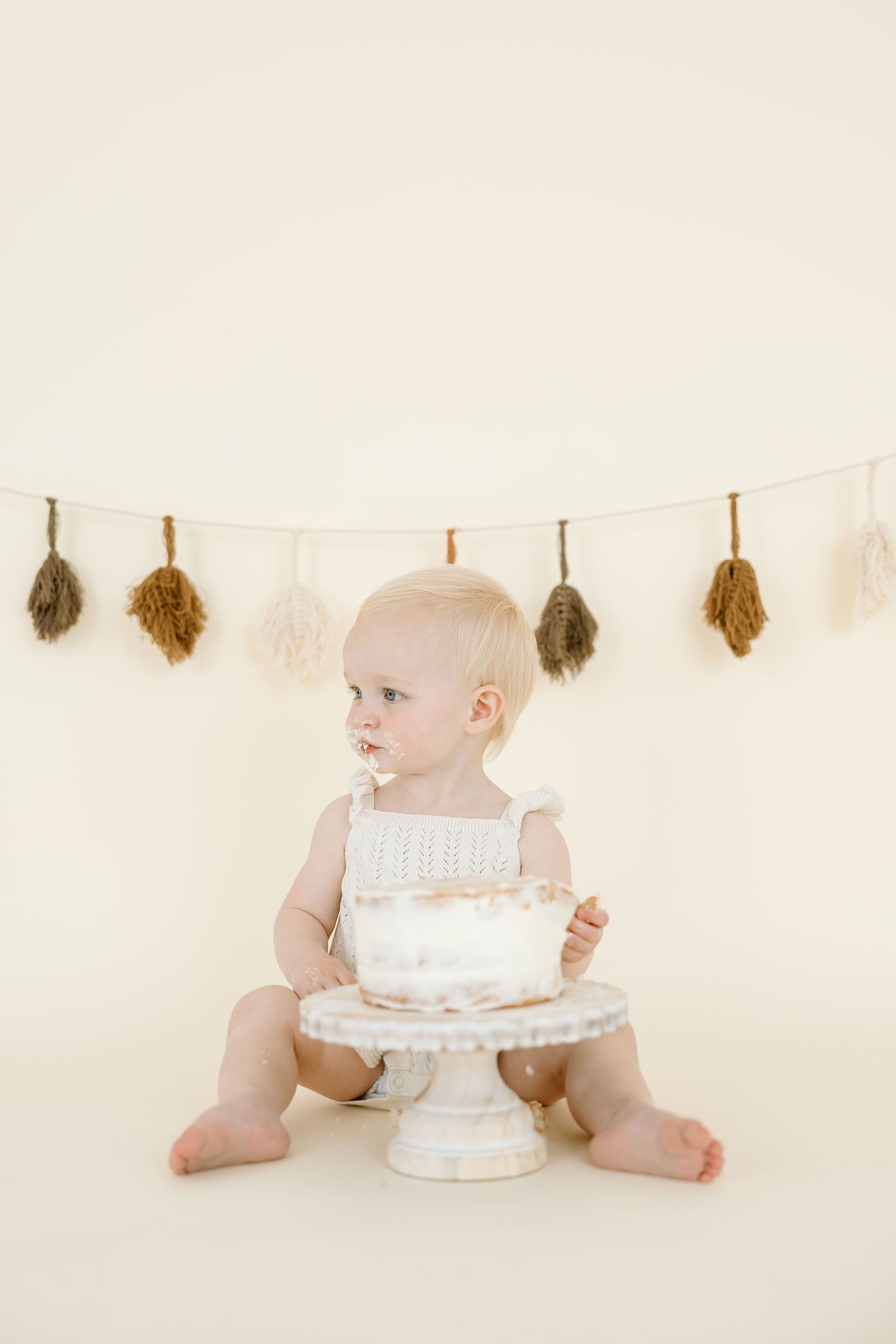 Baby in white outfit sits on cream backdrop with birthday cake and dried flower garland decorations.