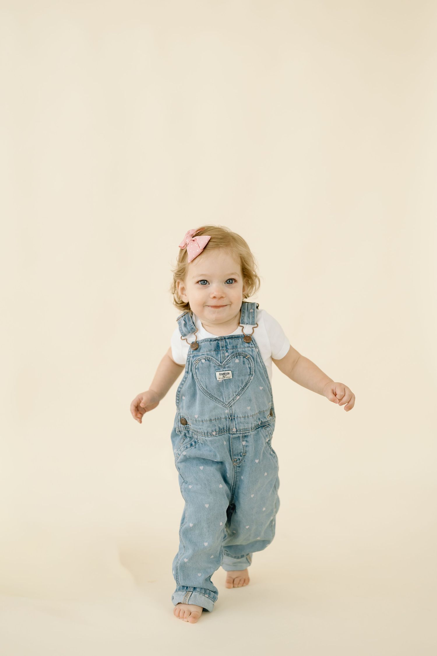 Small child wearing denim overalls and white shirt stands barefoot against neutral background.