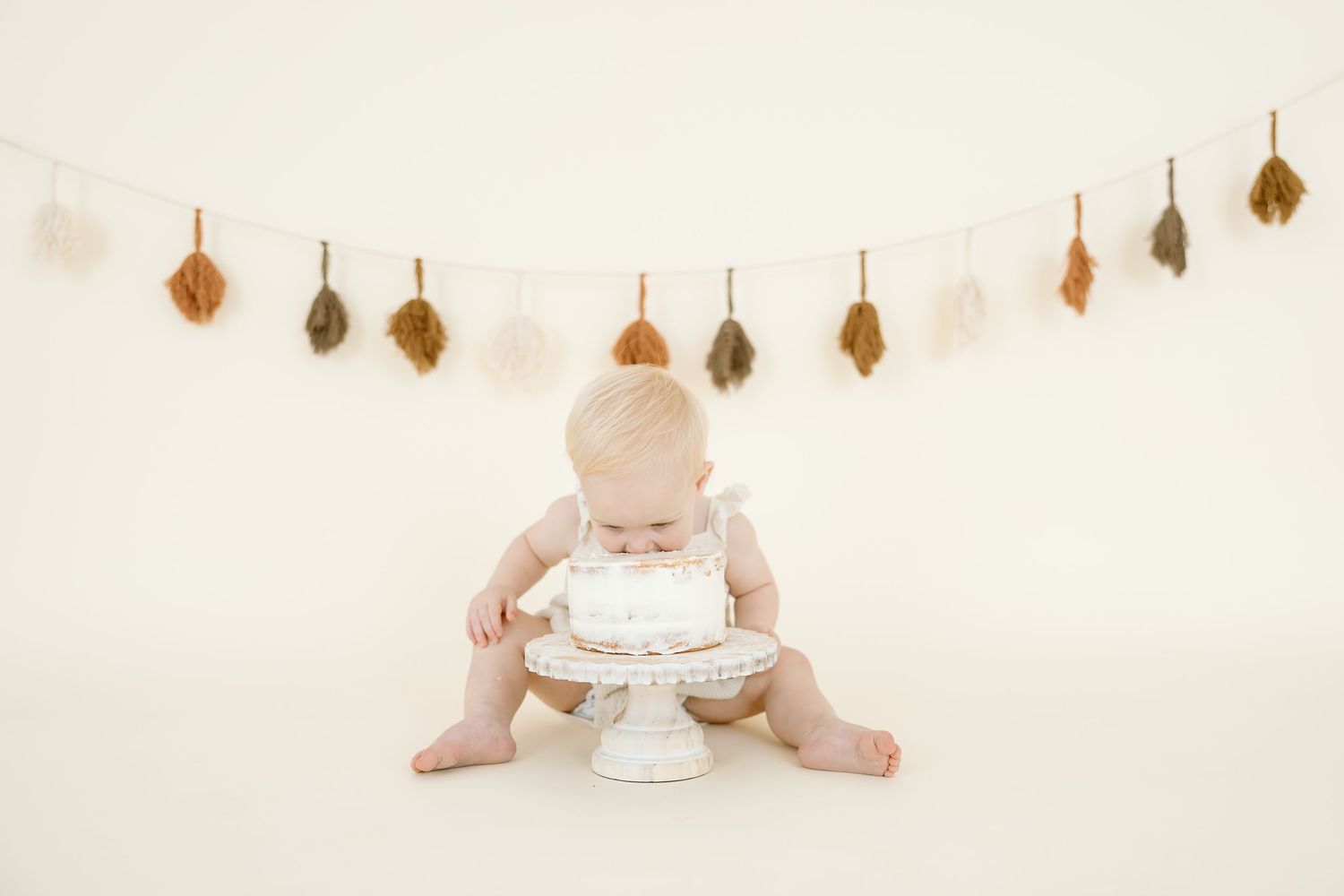 Baby in white outfit sits beside cake with gold leaf garland decorations hanging on cream wall behind.