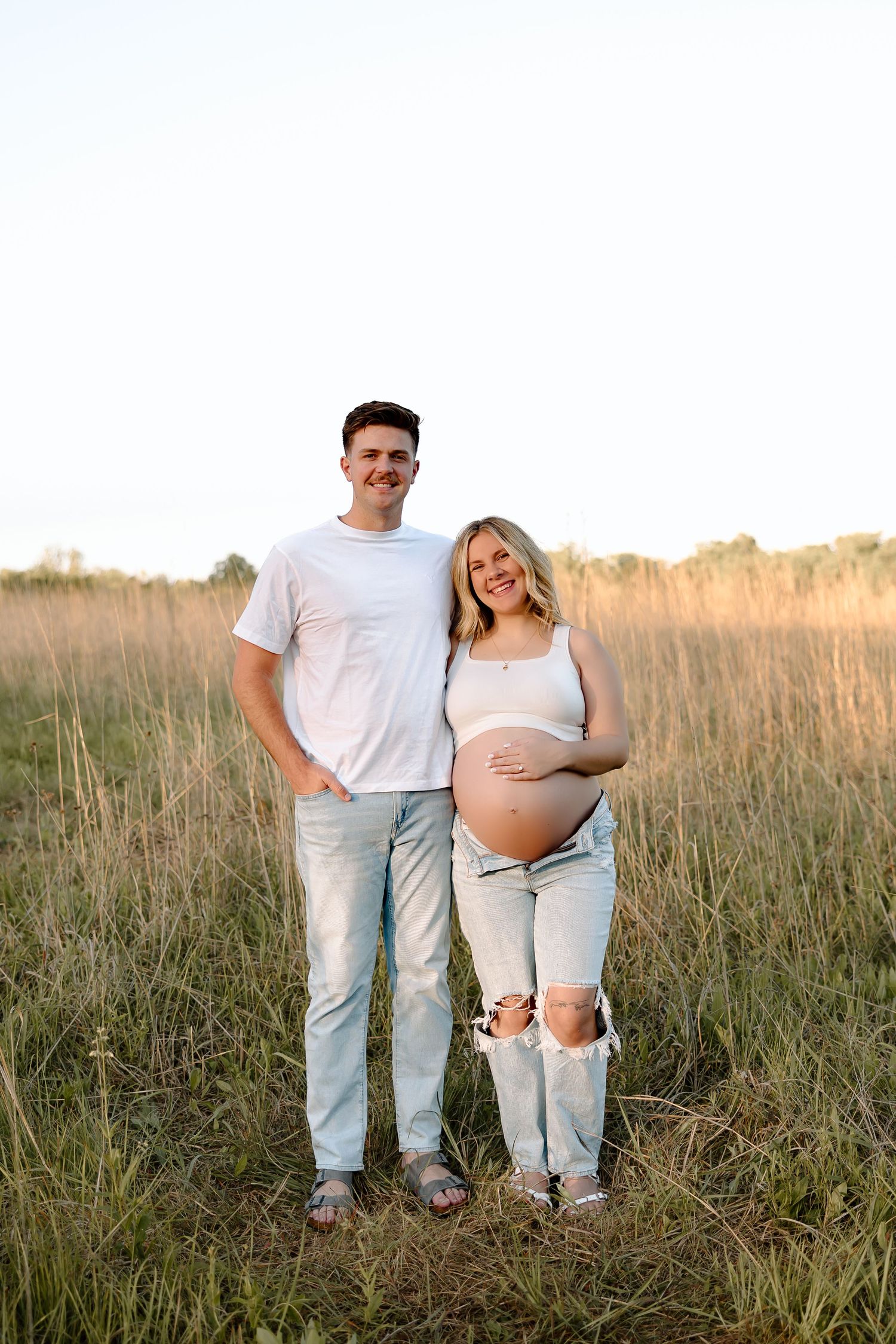 Expectant couple stands together in a sunlit meadow wearing casual white and denim outfits during golden hour.
