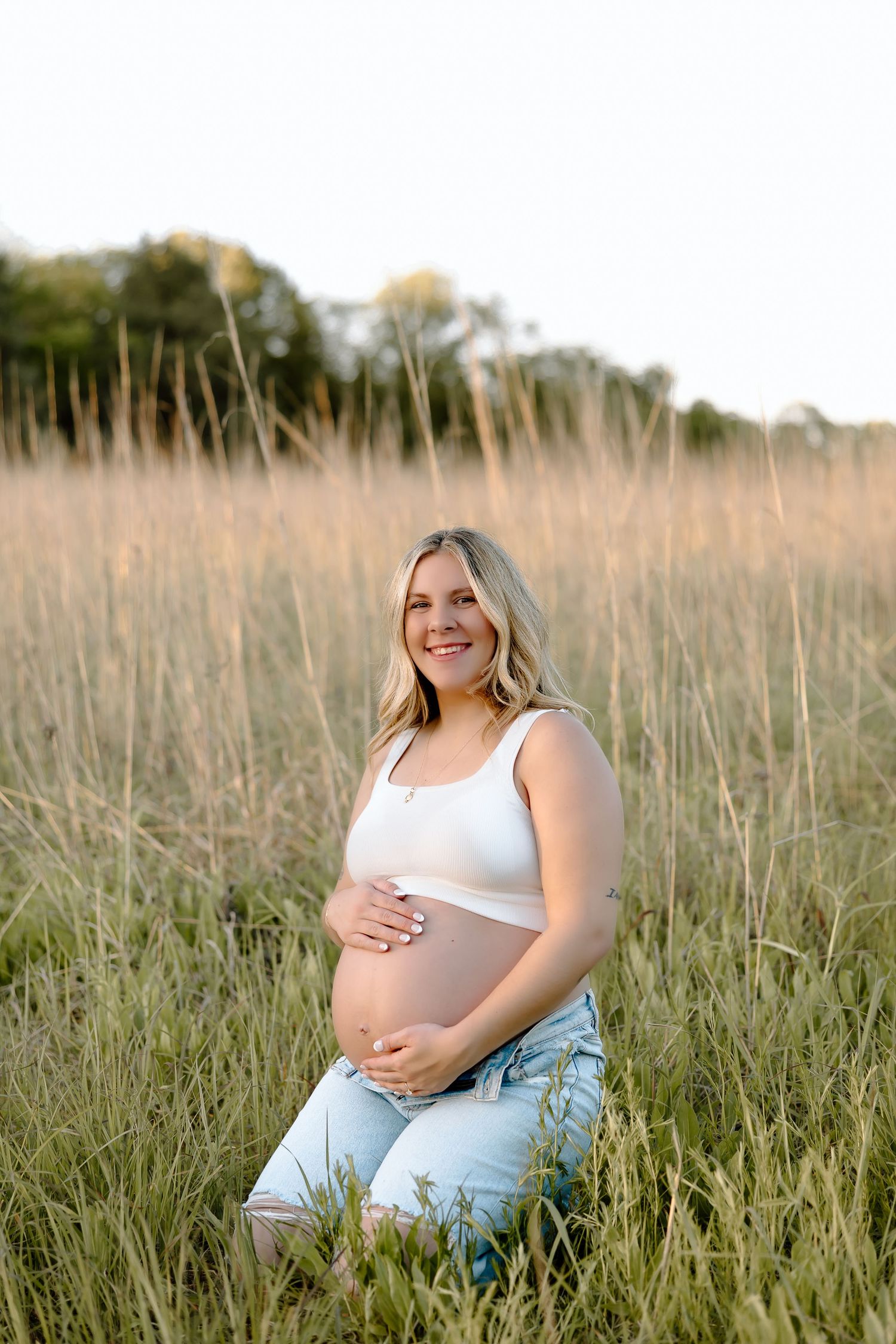 An expectant mother in a white tank top and jeans sits peacefully in a field of tall grass during golden hour.