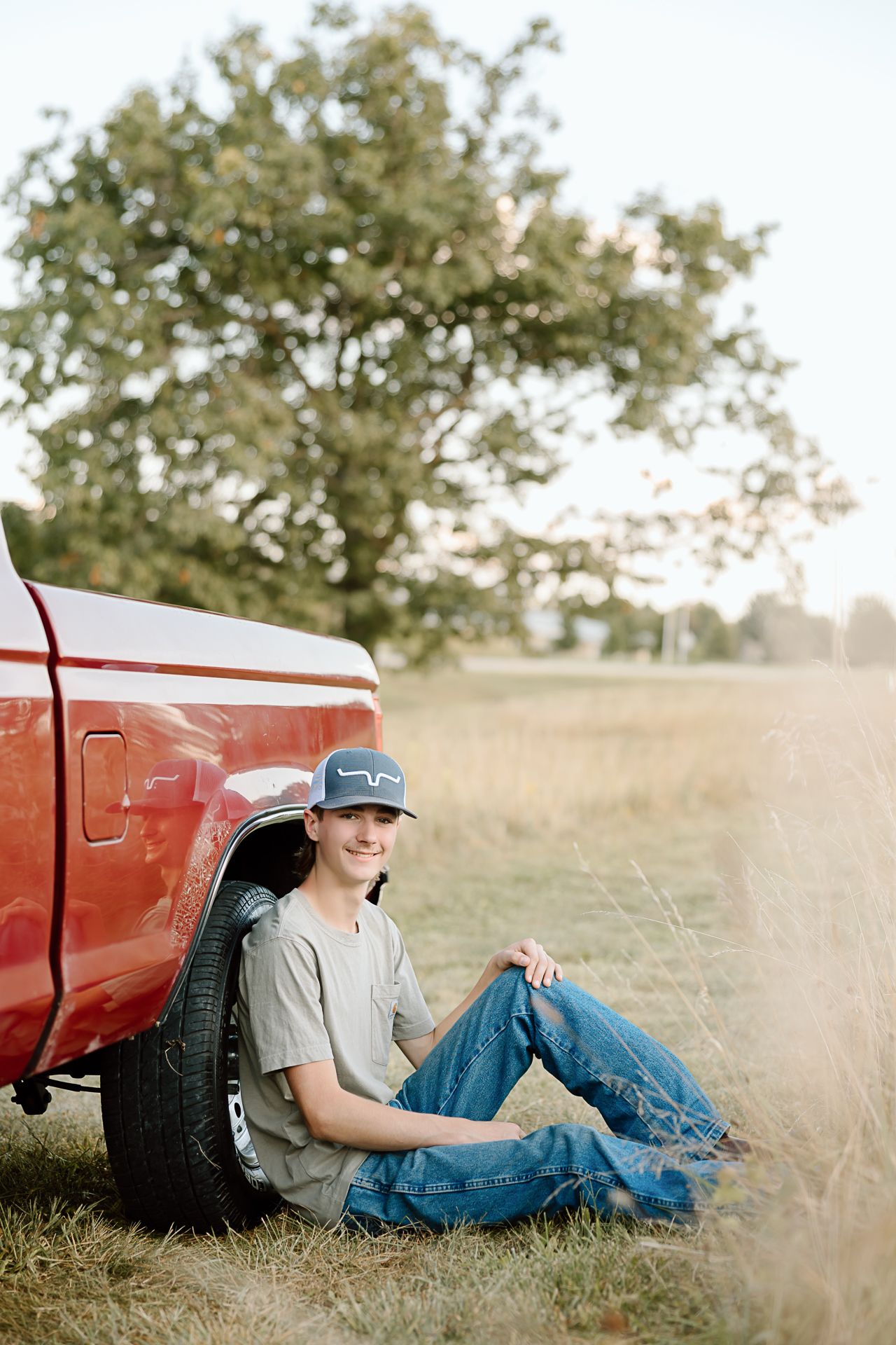 A couple relaxes in a grassy field next to a red pickup truck during a rustic countryside photoshoot.