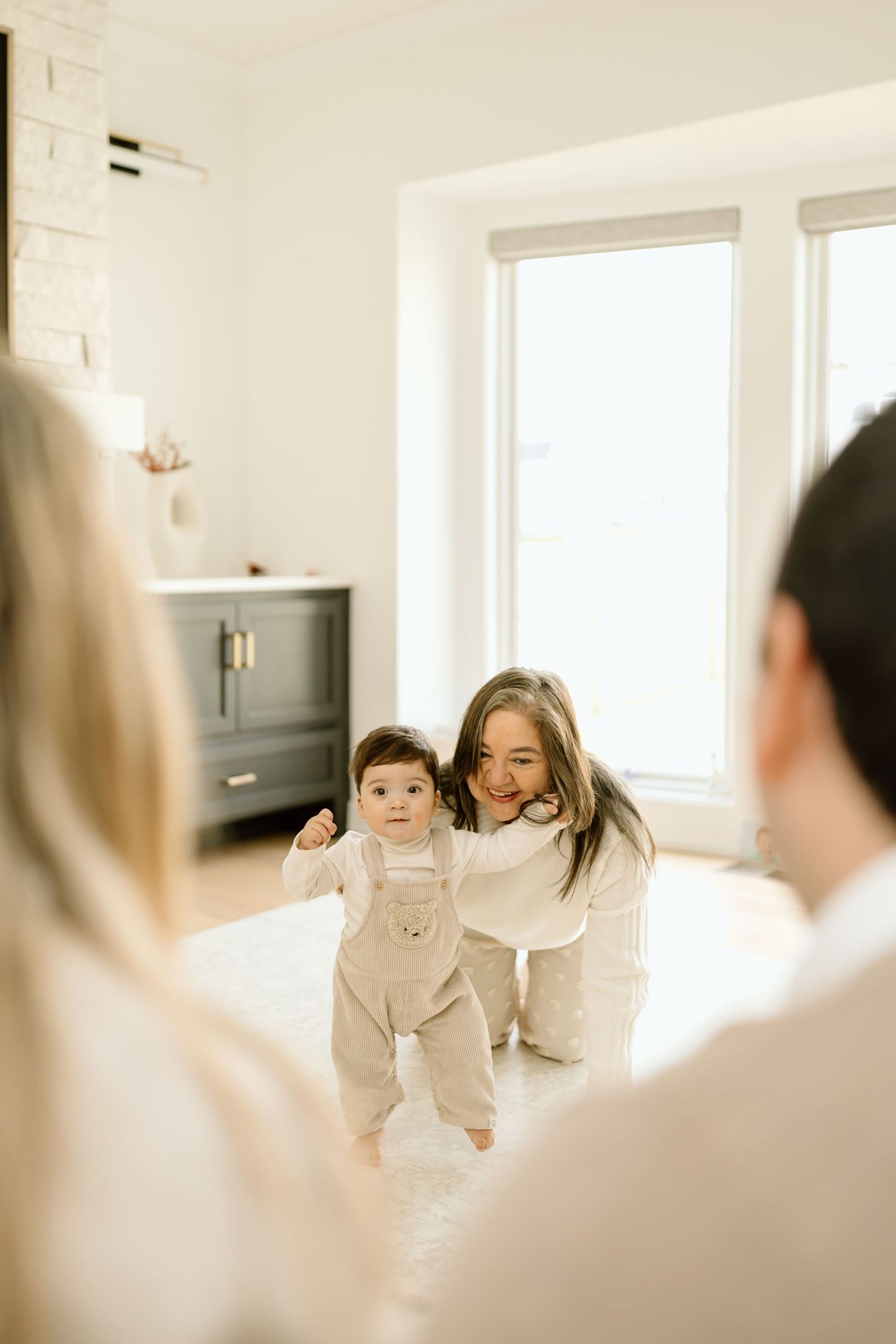 A bright interior room with white walls and large windows shows a tender family moment in soft, natural lighting.