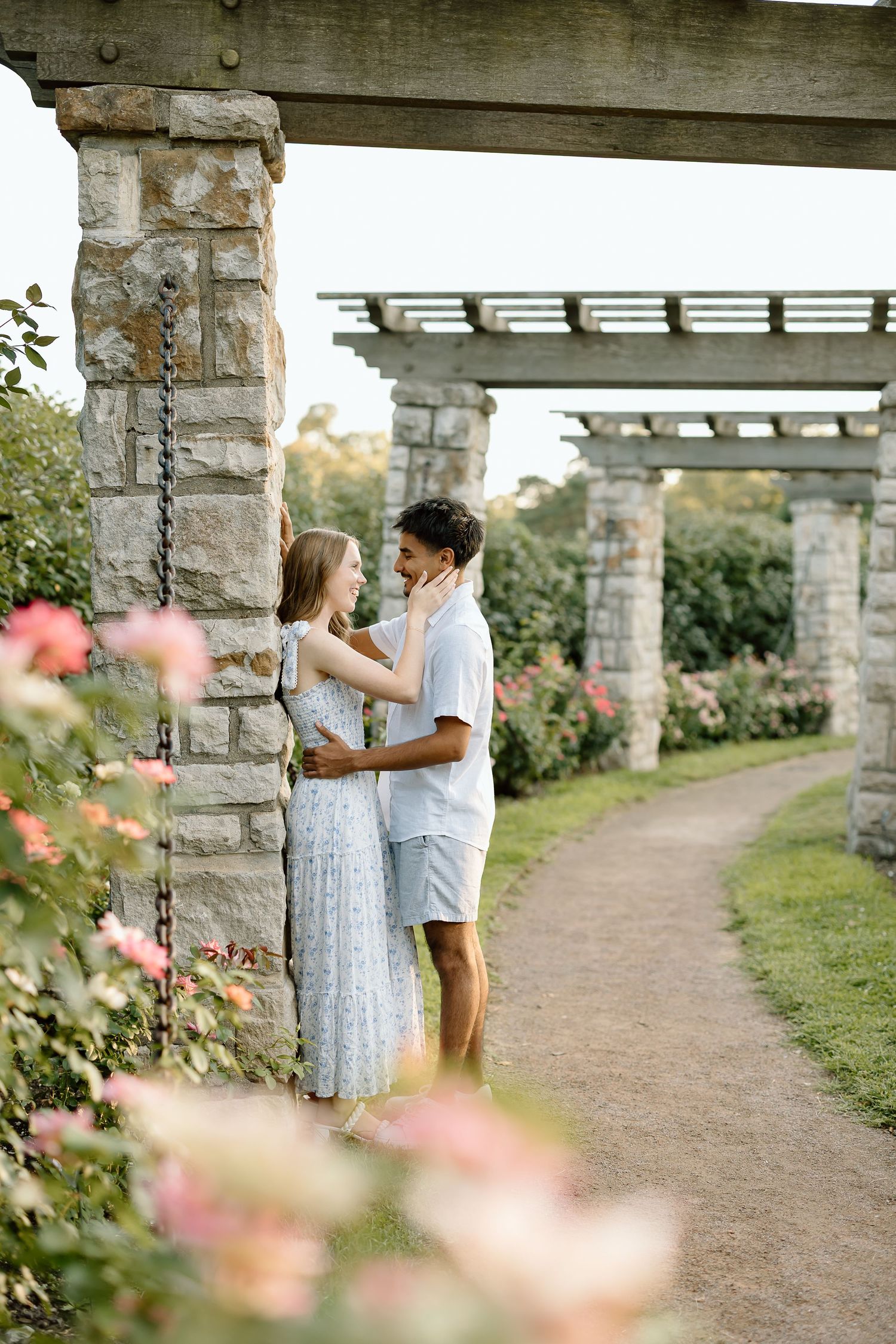 A couple shares a romantic moment under a stone pergola surrounded by blooming roses in a garden setting.