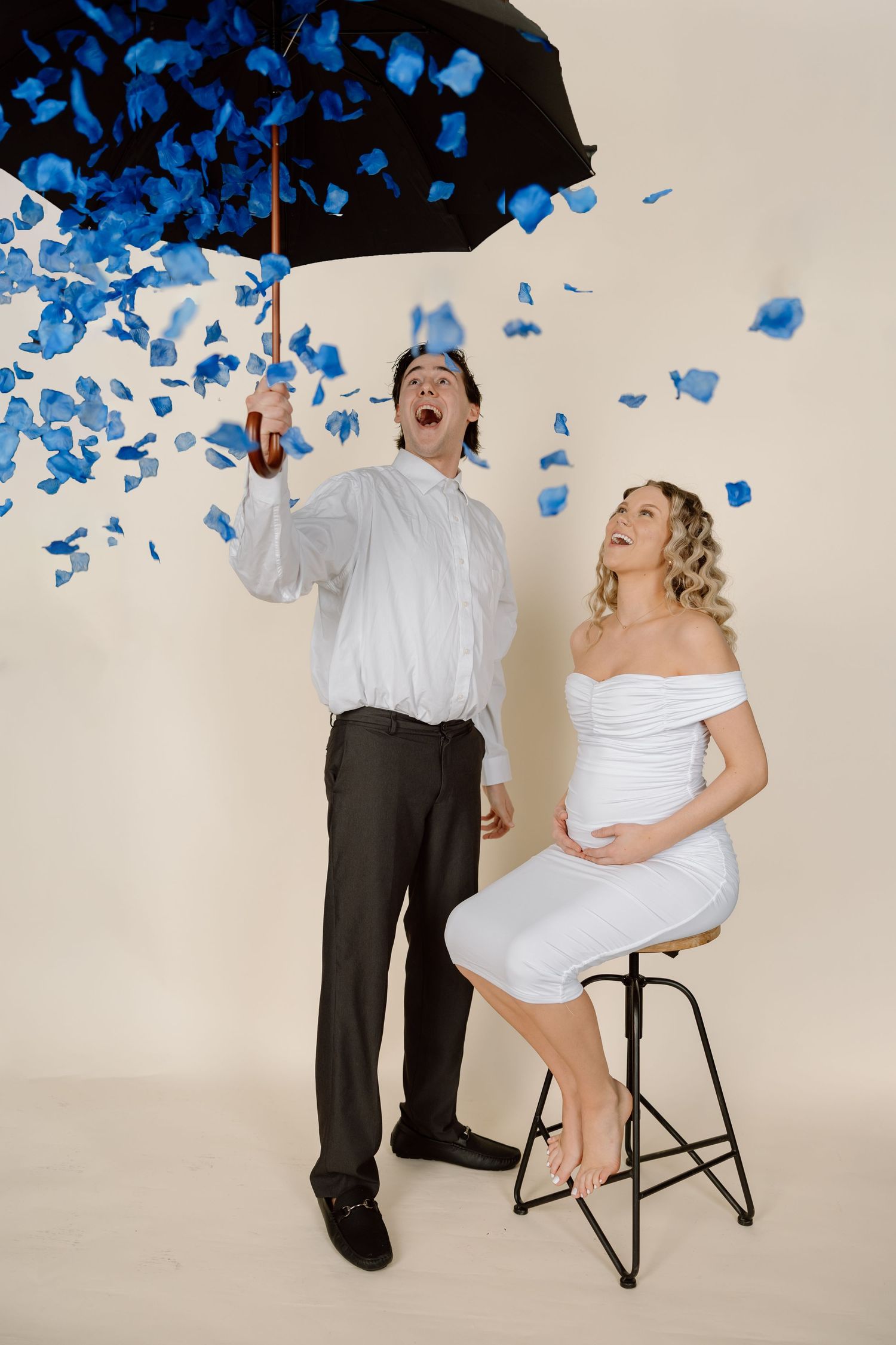 Couple in white attire poses playfully with blue confetti falling from above in a studio setting.