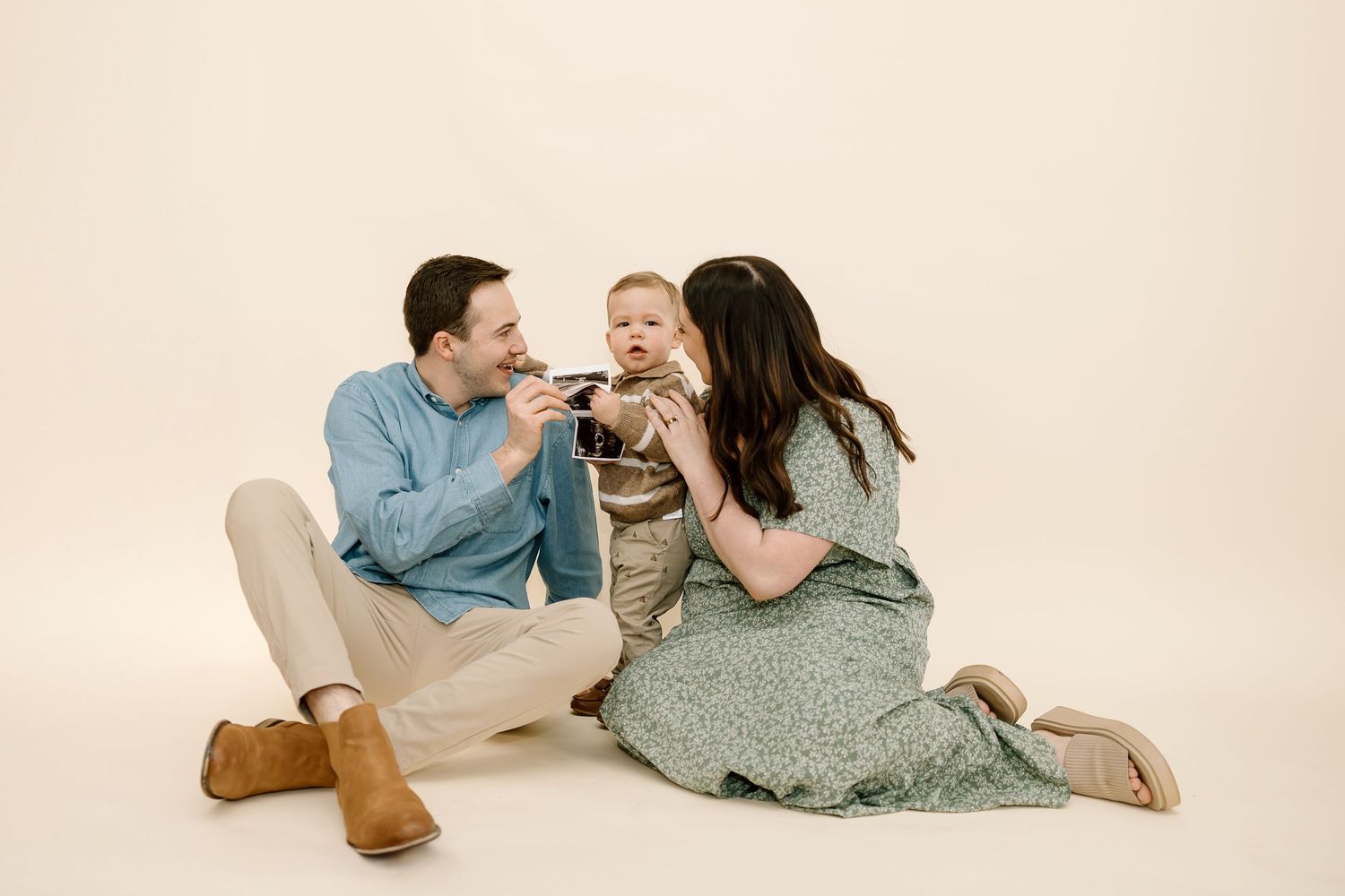 Family lying together on cream colored floor wearing casual clothing in soft natural light studio setting.