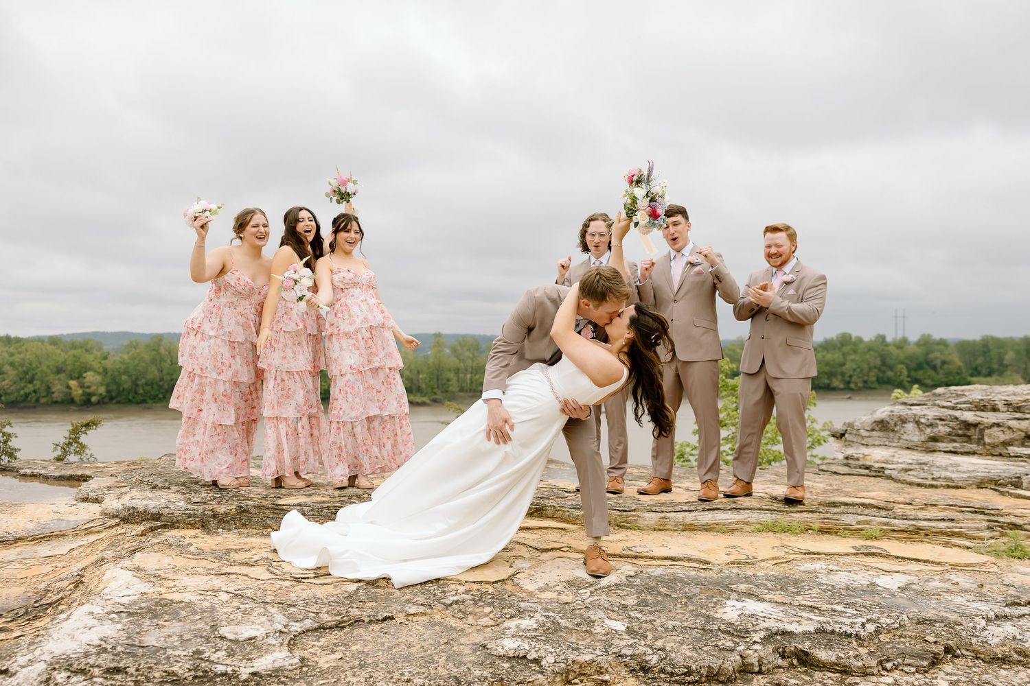 Wedding party in pink dresses and tan suits pose together on a scenic rocky overlook with mountain views in the background.