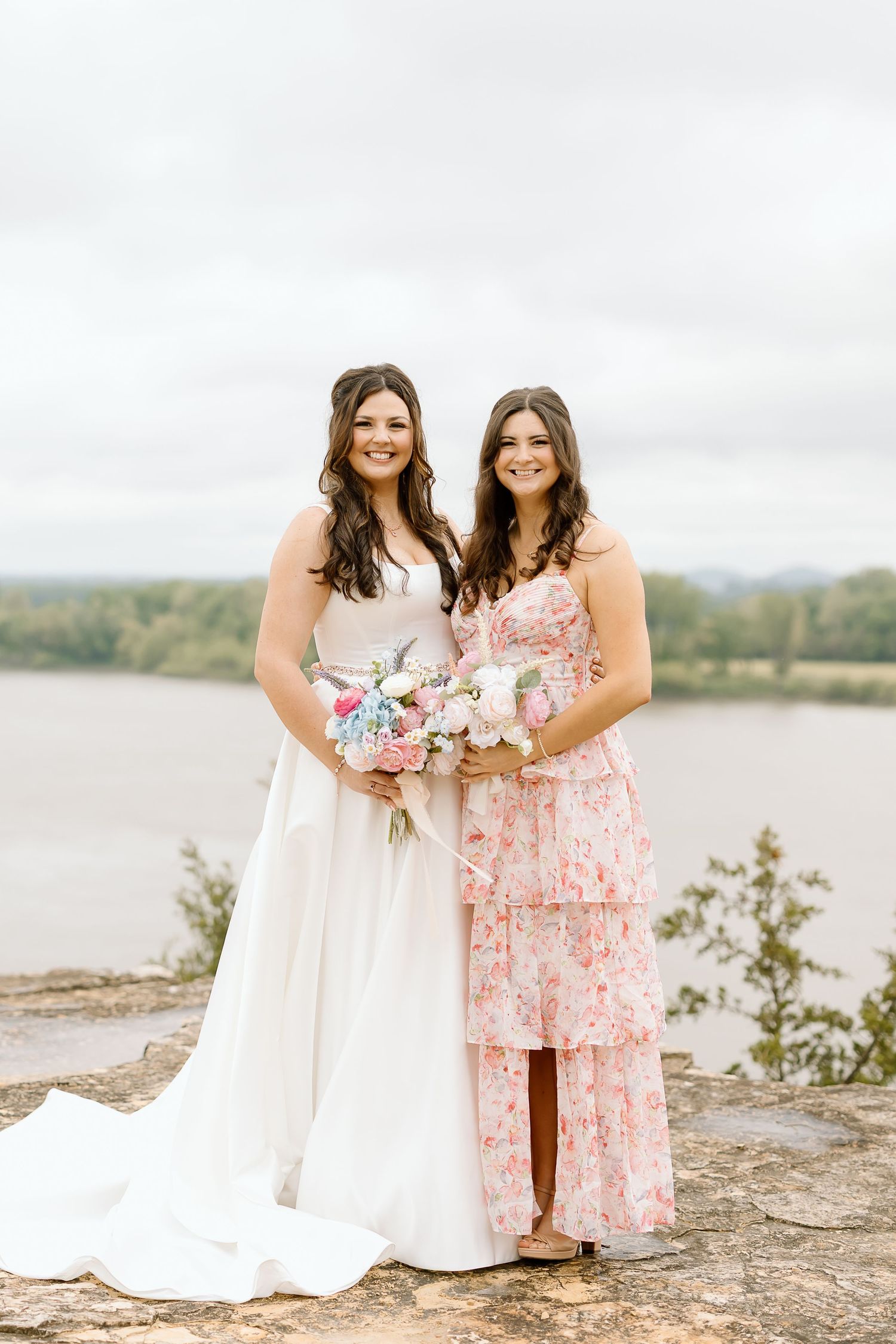 Two women in white and floral pink dresses pose together by a scenic lakeside on rocky terrain.