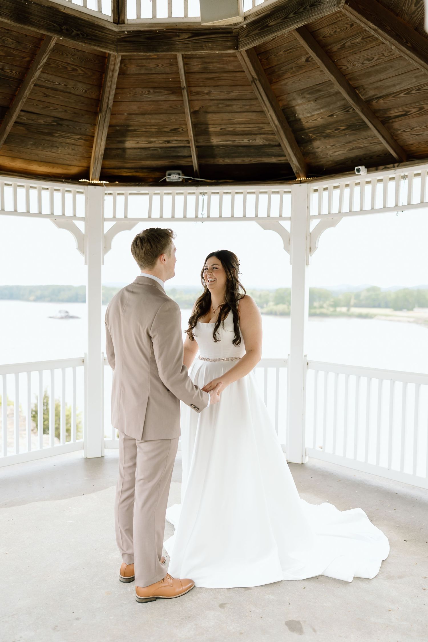 A romantic wedding couple shares an intimate moment in a white wooden gazebo overlooking a waterfront view.