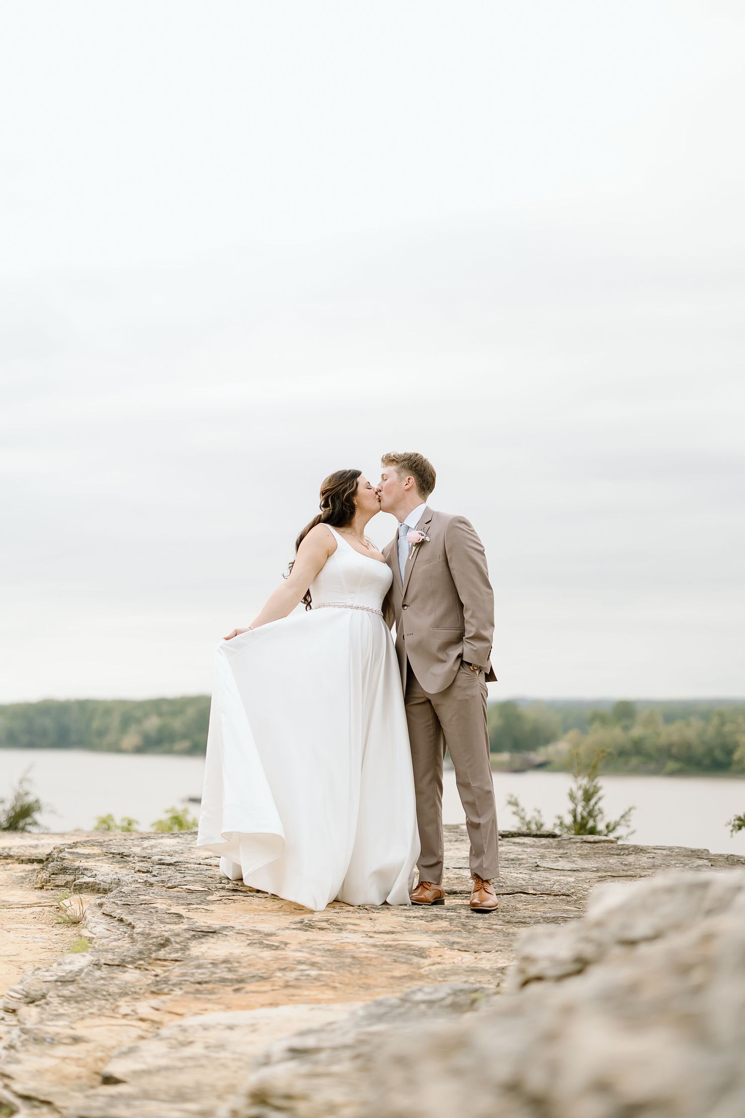 Romantic wedding couple embraces on rocky cliff overlooking scenic lake during sunset photoshoot.