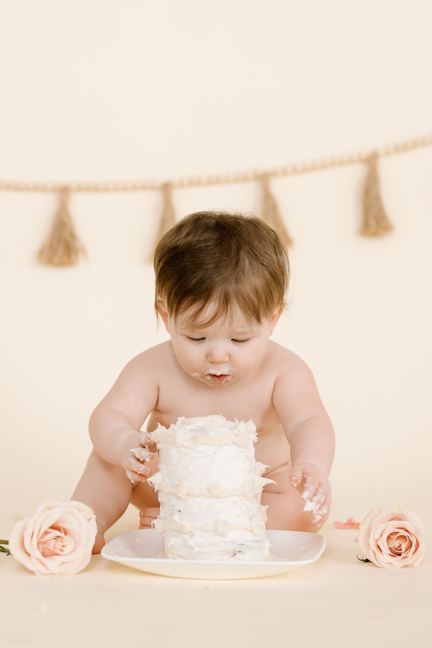 Curious baby exploring white frosted cake with messy hands beneath hanging tassel garland.