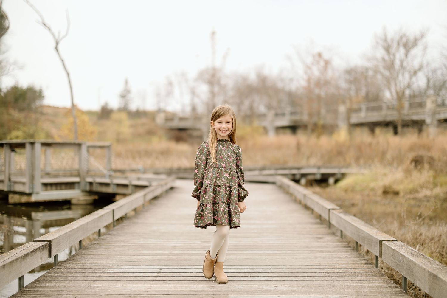 A child poses while standing on a wooden boardwalk surrounded by autumn scenery.
