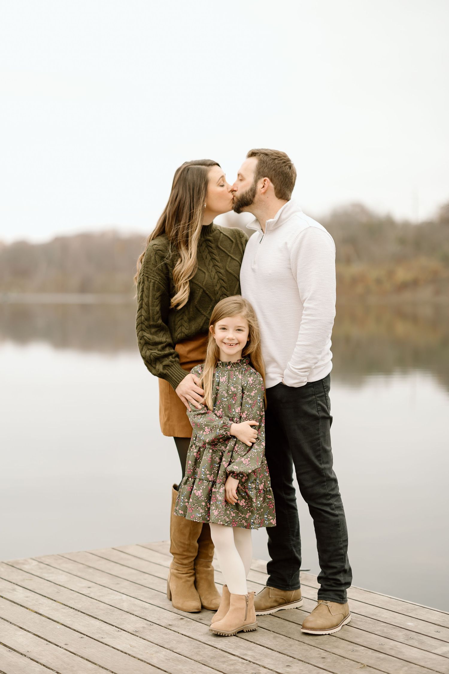 Family of three embraces while standing on lakeside dock in coordinated fall outfits.