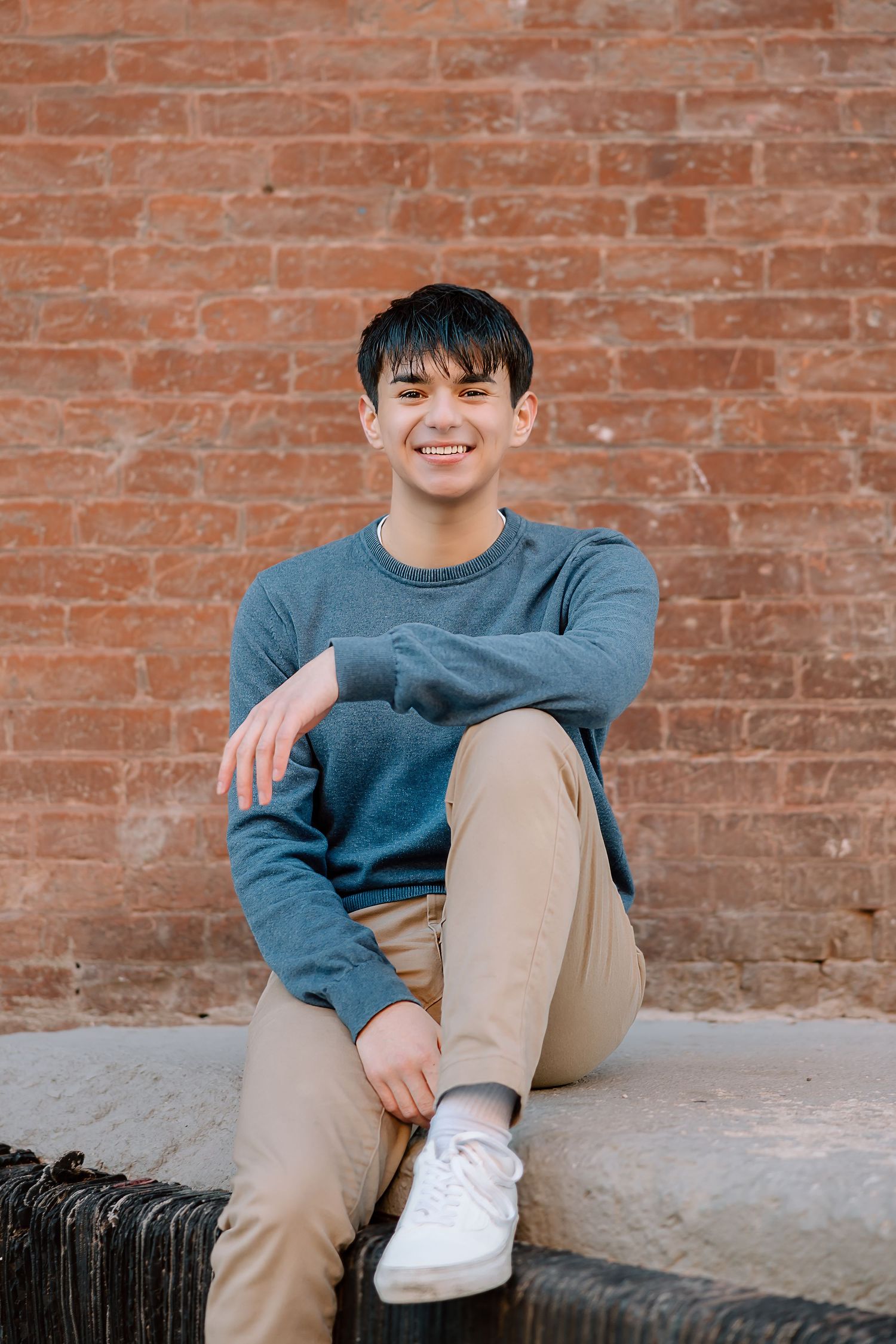 Casual portrait sitting on concrete ledge against brick wall in blue sweater and khaki pants.