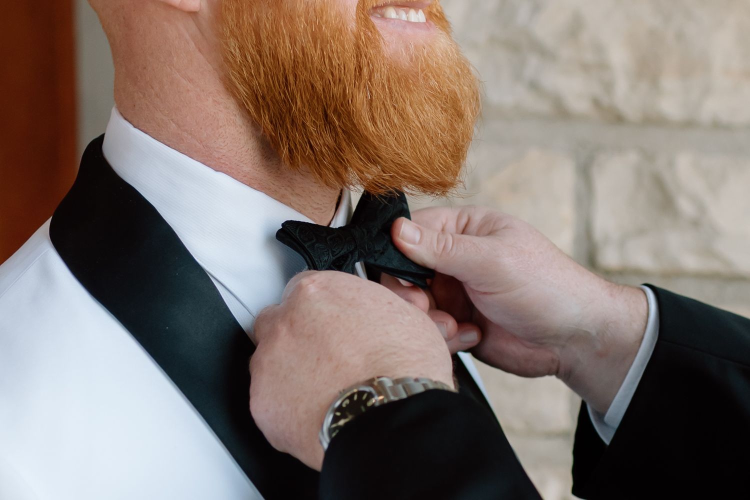 A ginger-bearded person wearing a black suit adjusts their tie in a series of close-up shots against a stone wall.