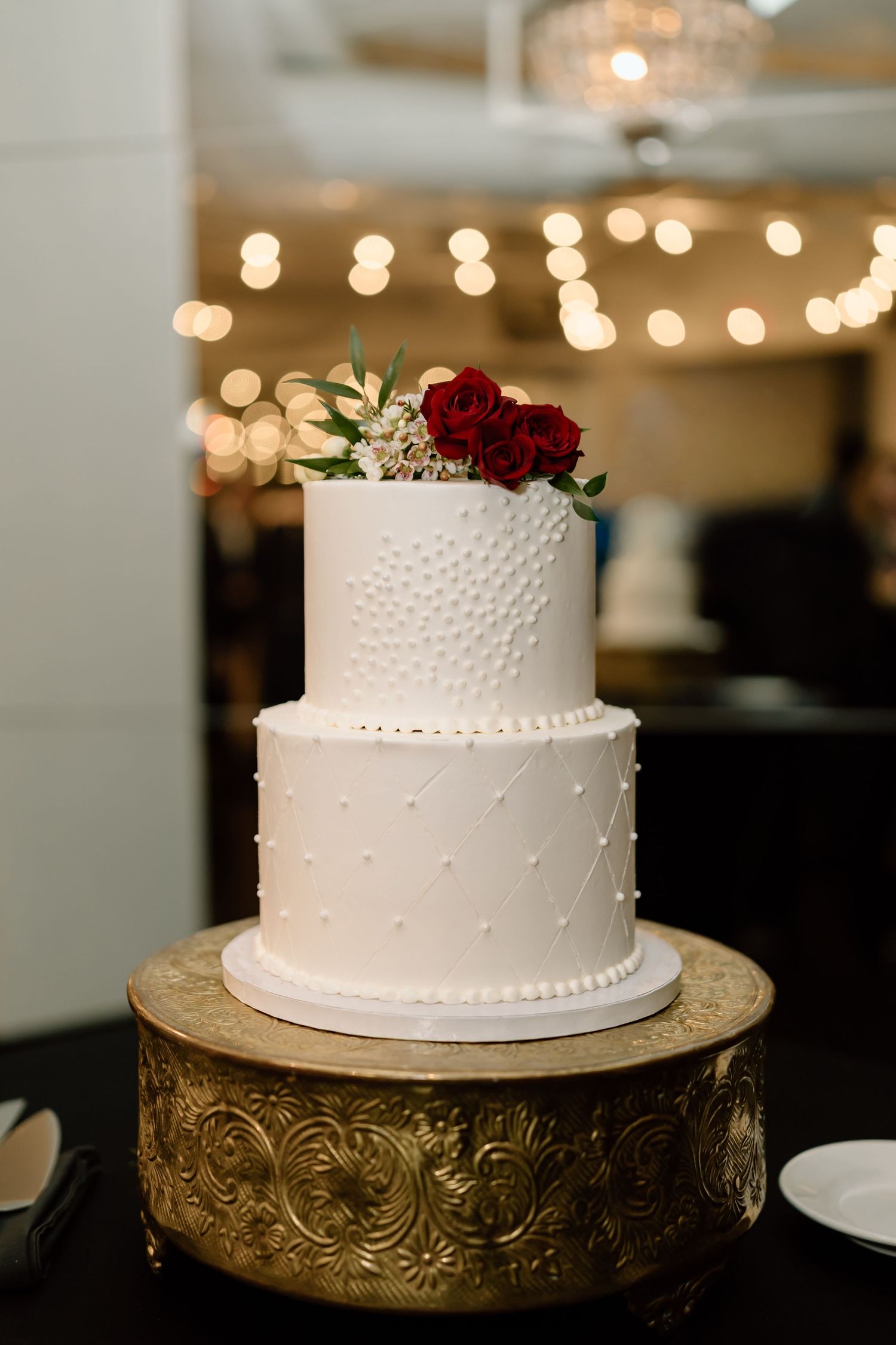 Elegant two-tier white wedding cake with textured dots and red roses on ornate gold stand with bokeh lights in background.
