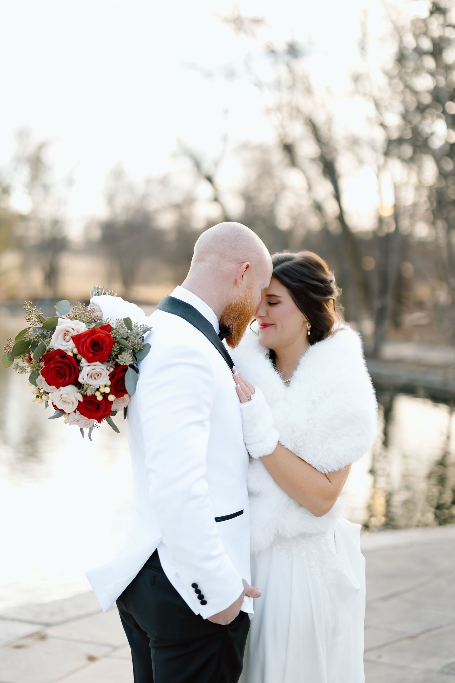 Romantic winter wedding couple in white embraces outdoors with red bouquet at sunset.