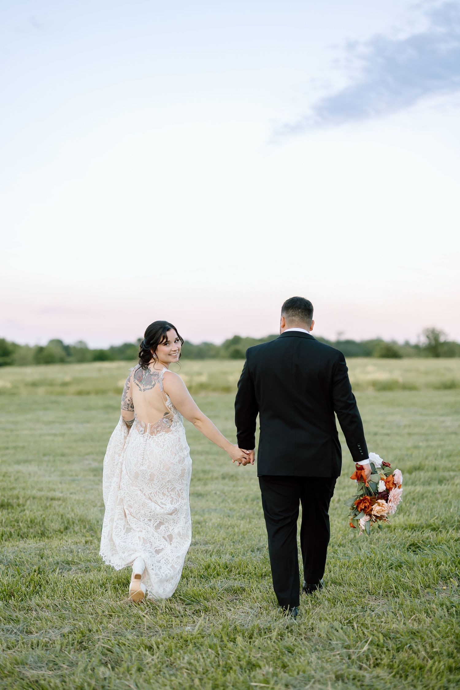 A bride in a lace dress and groom in black suit walk together across a green meadow holding a vibrant floral bouquet.