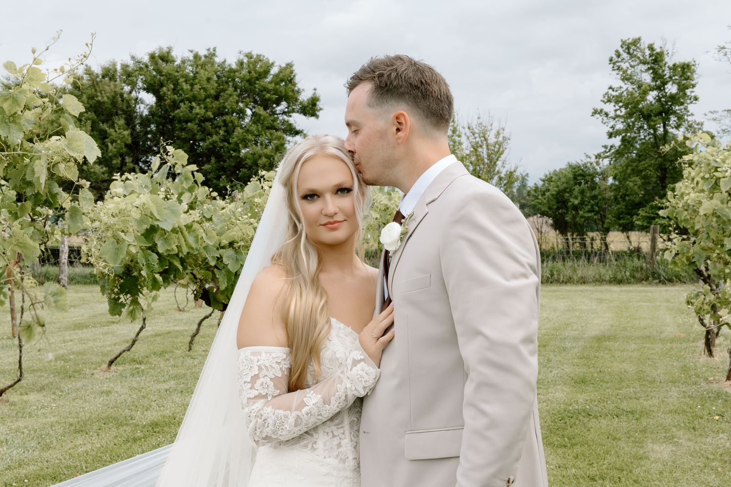 Groom in beige suit kisses bride in lace off-shoulder gown and veil outdoors at vineyard wedding.