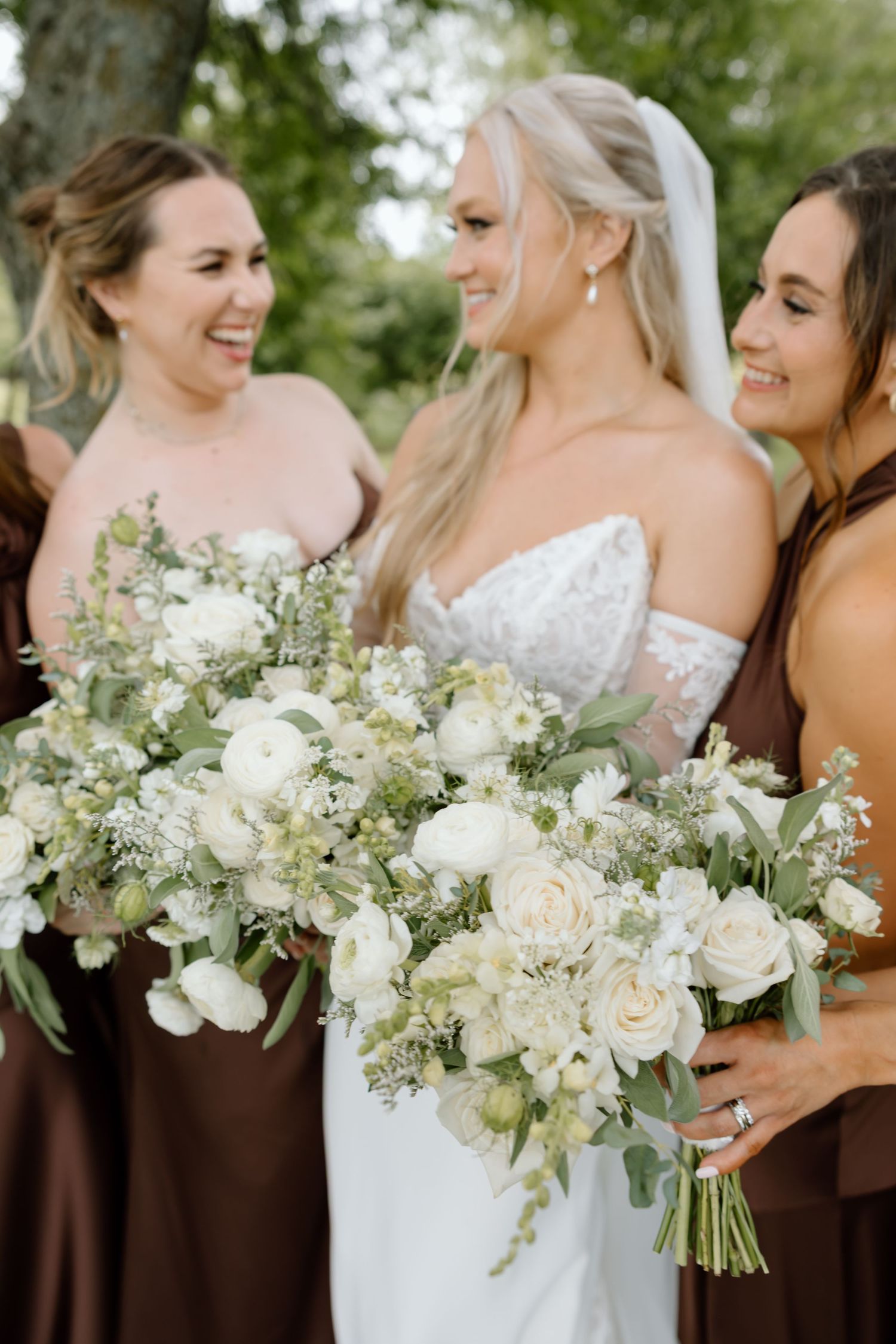 Bride in white lace gown holds bouquet with bridesmaids in brown dresses, all carrying white floral arrangements.