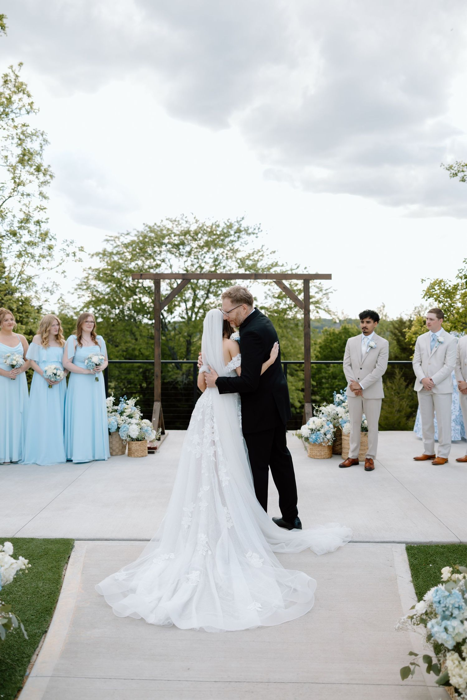 Bride and groom share first kiss at outdoor wedding ceremony with blue floral arrangements and wedding party.