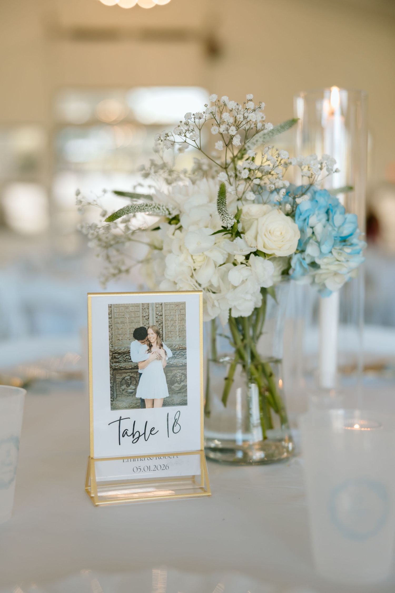 Elegant wedding table centerpiece with white and blue hydrangeas, roses, and a Polaroid Table 18 card in a gold frame.