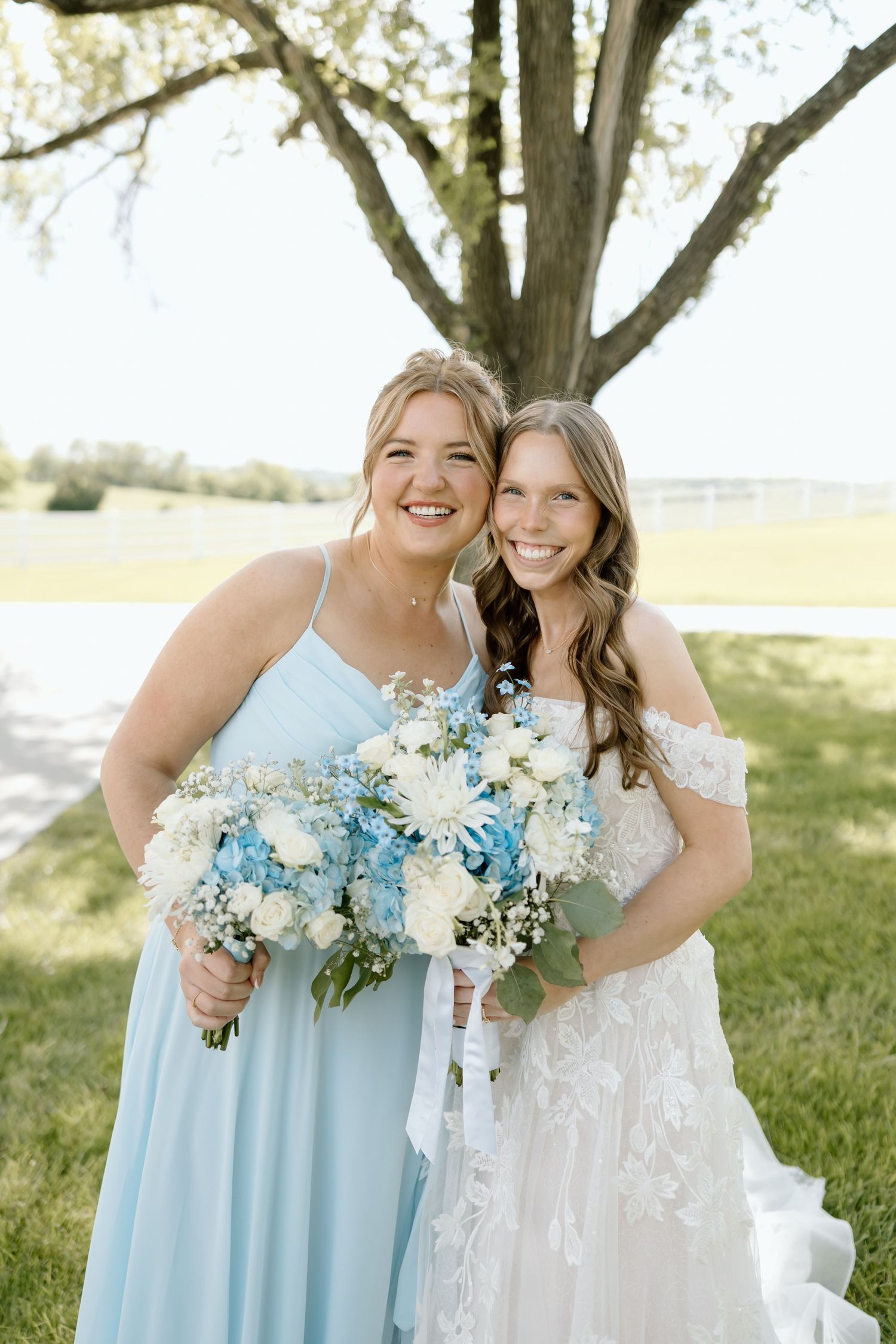 Smiling bride and bridesmaid in blue dress hold white and blue bouquets under a tree.