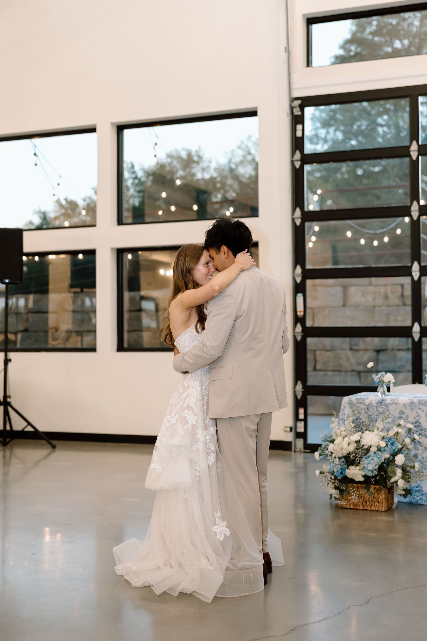 A bride and groom share their first dance in an elegant venue with string lights and large windows in the background.