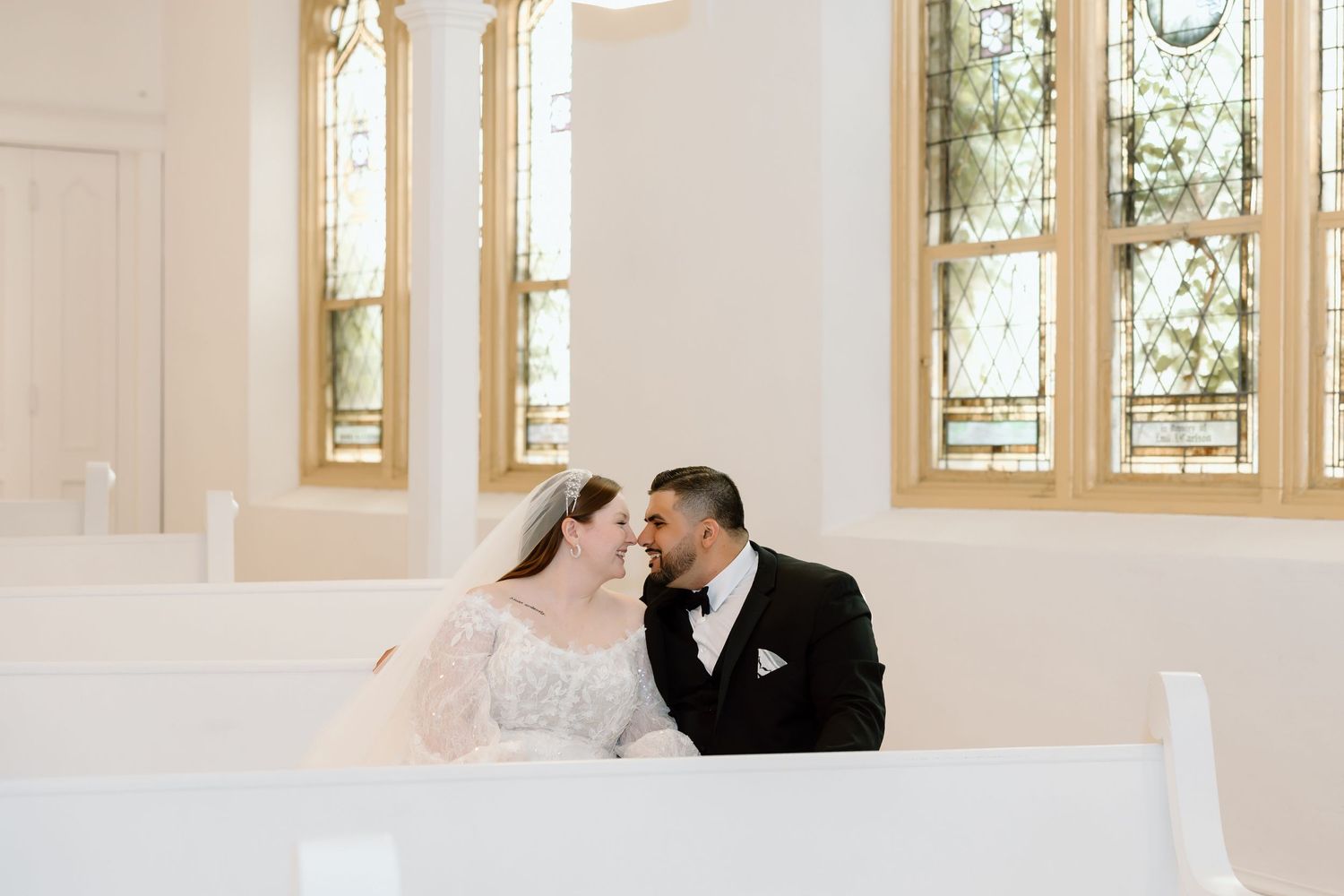 Bride and groom sitting together in a white church pew with golden stained glass windows in the background.