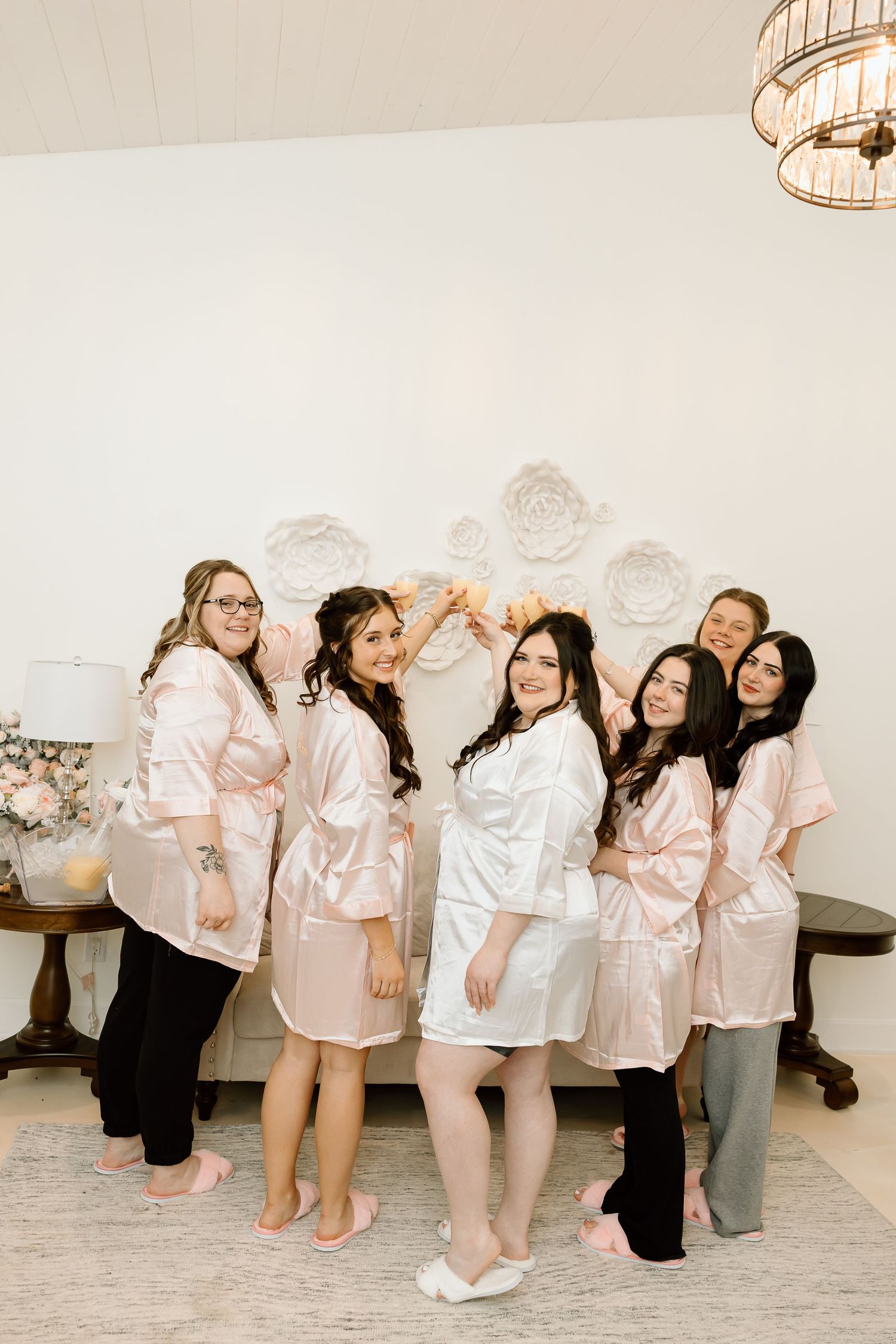 A group in matching pink and white satin robes poses together in a room with decorative wall details and lighting.