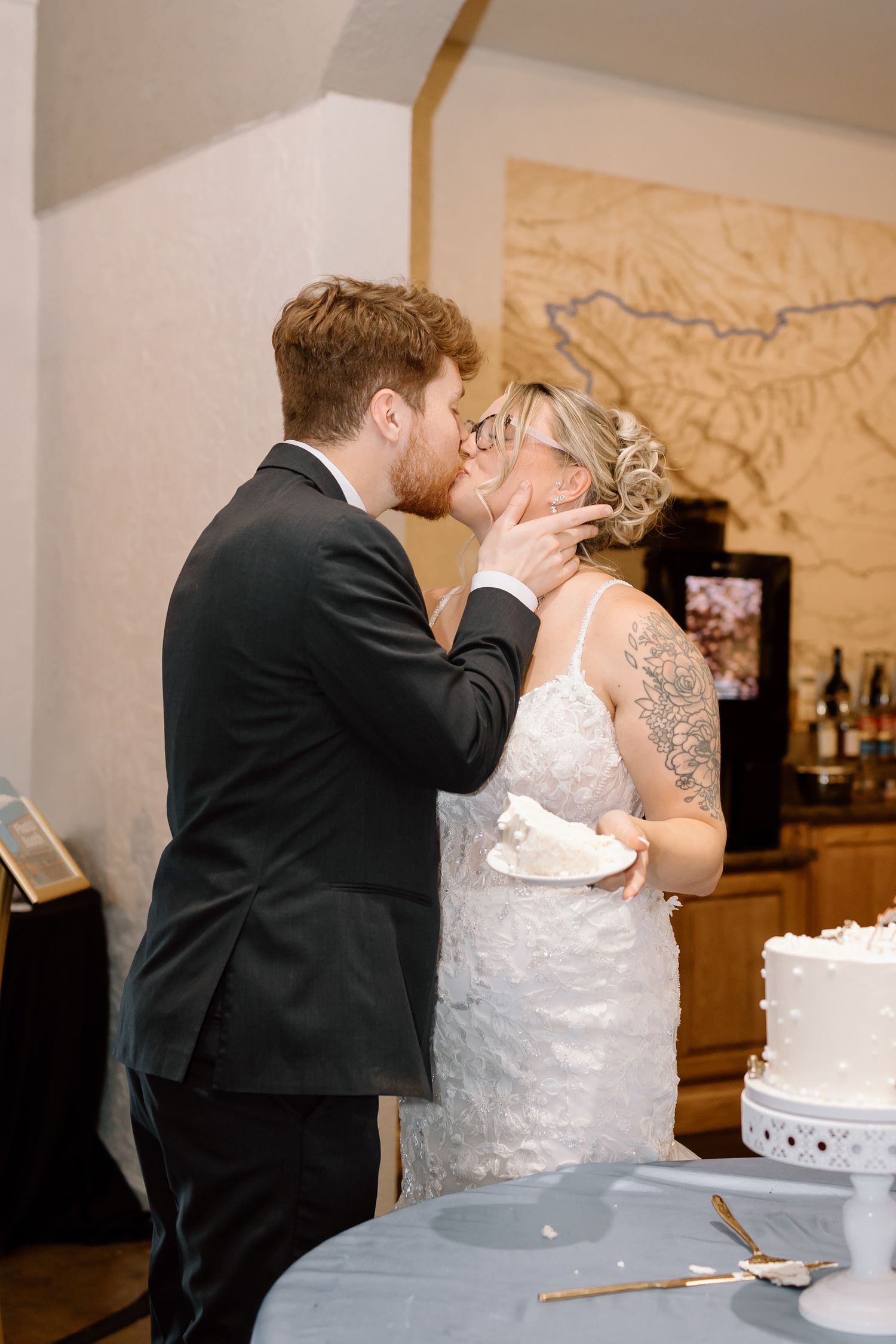 A bride and groom share their first kiss as newlyweds next to their wedding cake at the reception.