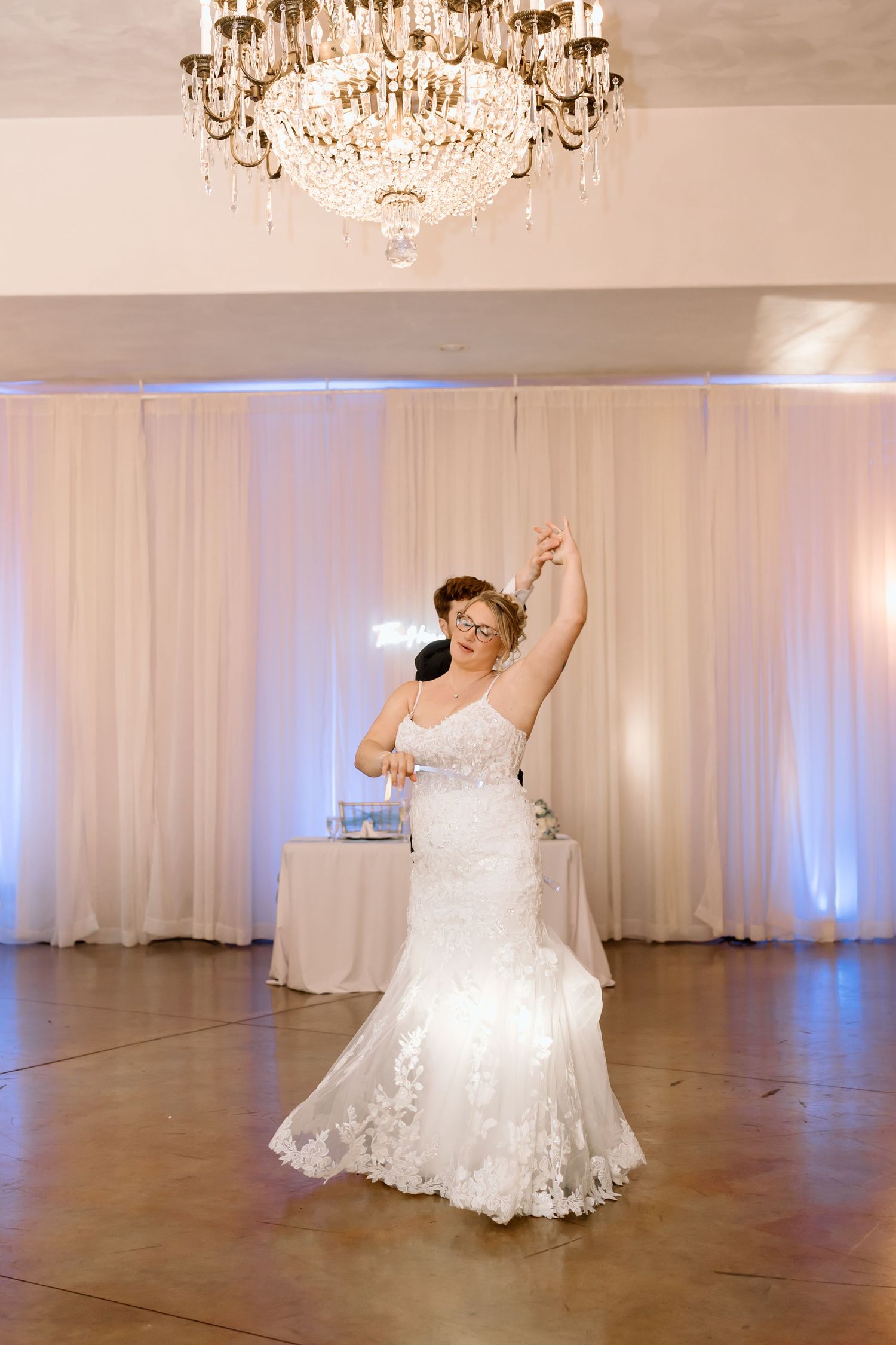 A bride in a white lace gown twirls on a polished dance floor under crystal chandeliers.