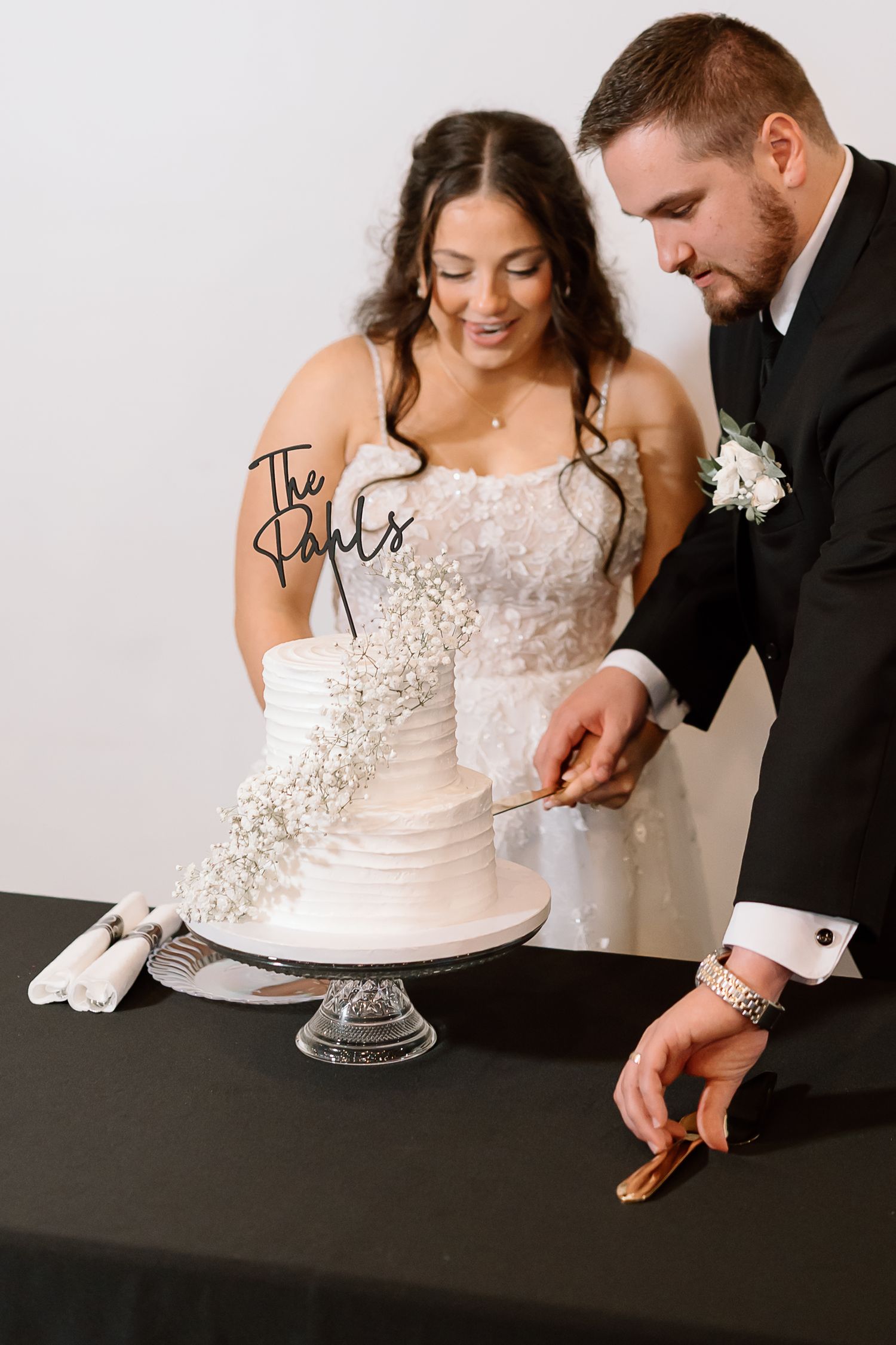 A couple cuts into their elegant white wedding cake adorned with delicate floral details on a clear glass cake stand.