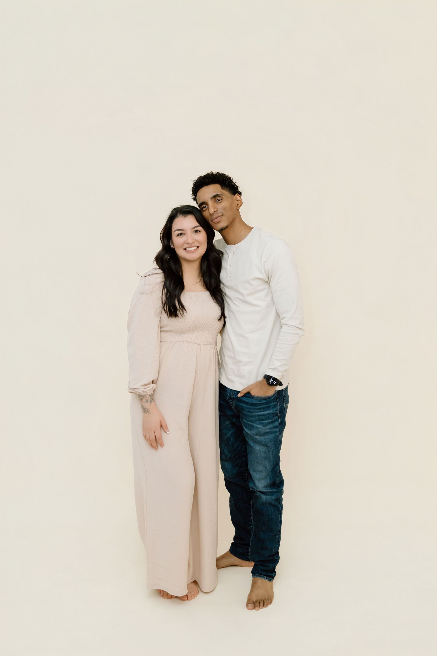 A couple in coordinating neutral outfits poses together against a light beige backdrop in a minimalist studio setting.