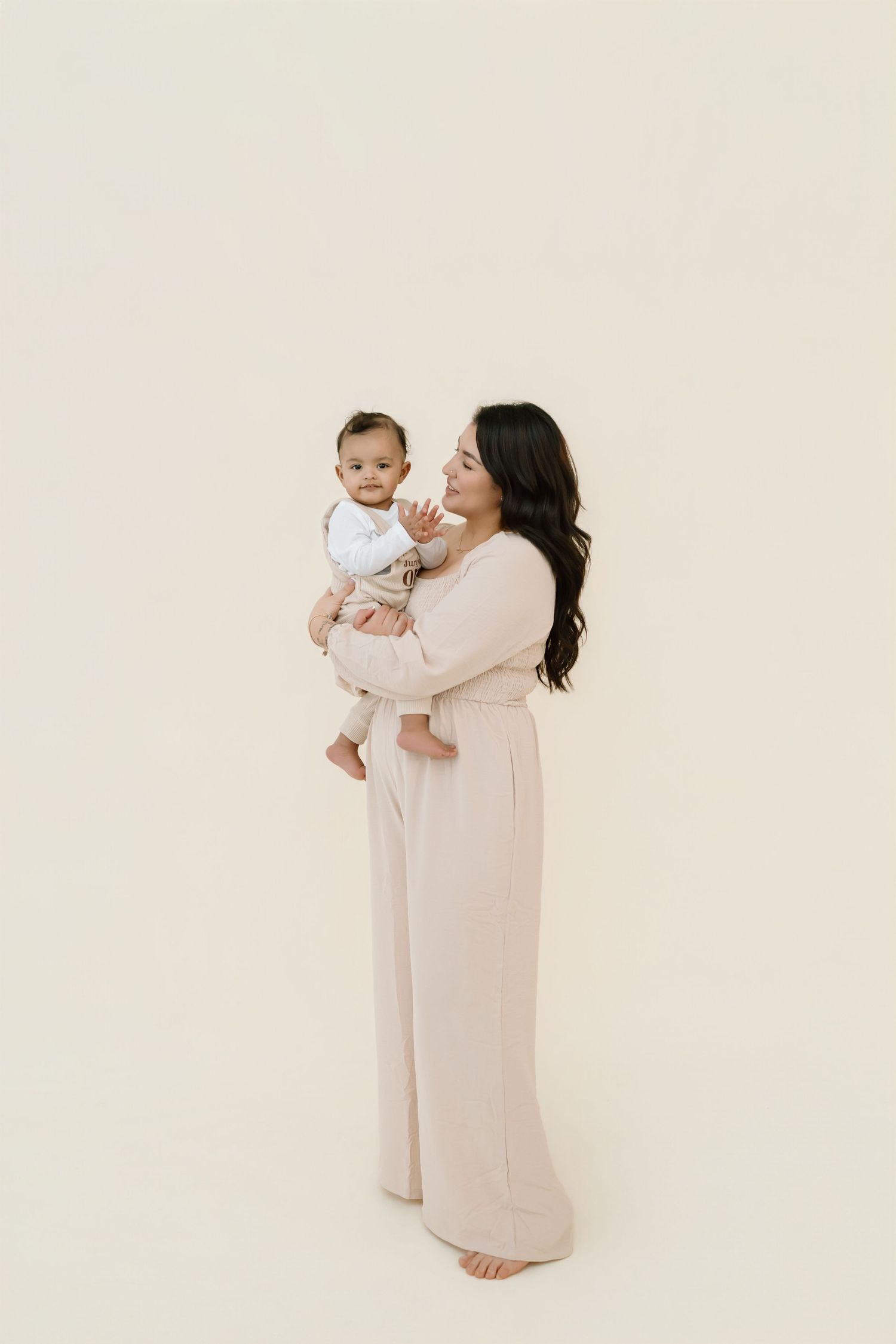 Parent in cream-colored dress holds baby against neutral background in minimalist studio portrait series.