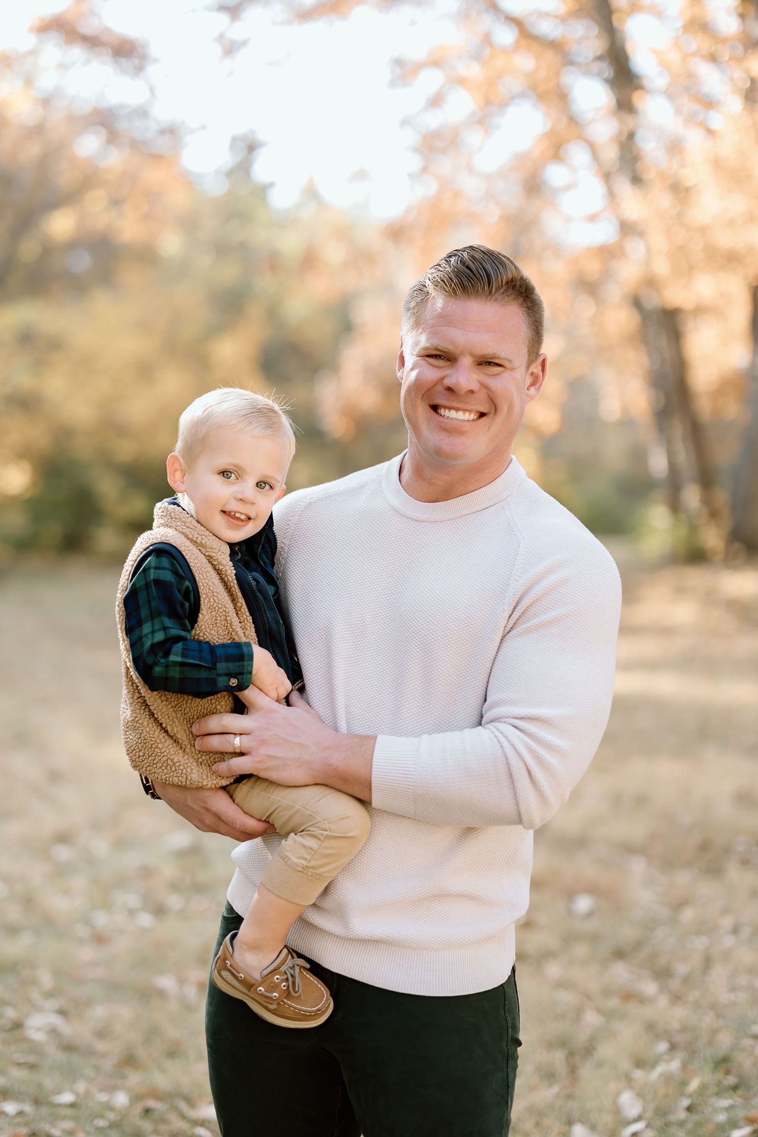 A parent in a white sweater holds a smiling child wearing plaid and tan during an autumn photoshoot.