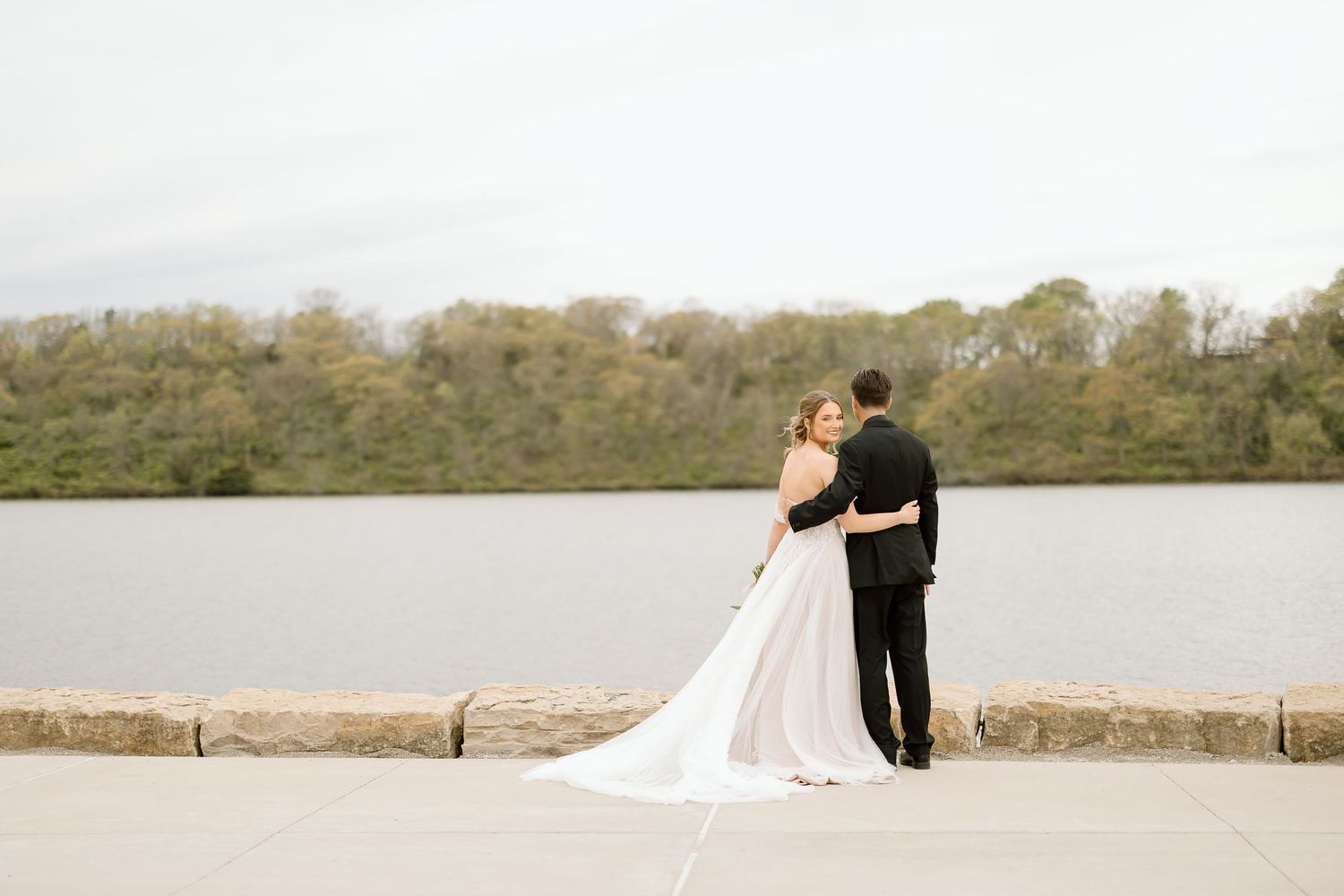 Couple shares intimate moment by lakeside stone wall during wedding photoshoot.