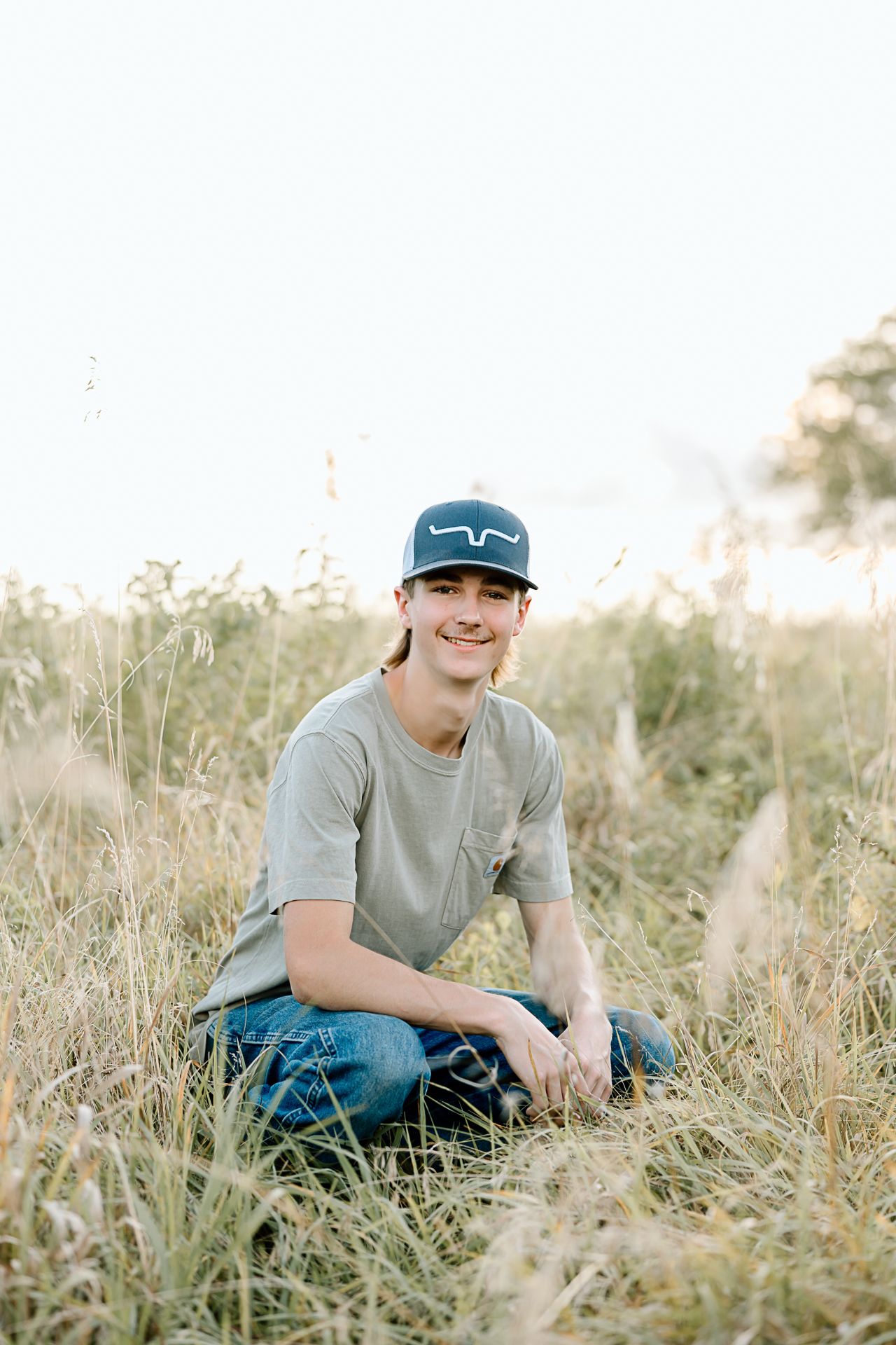 Person sitting in tall grass wearing blue cap and gray t-shirt during golden hour.