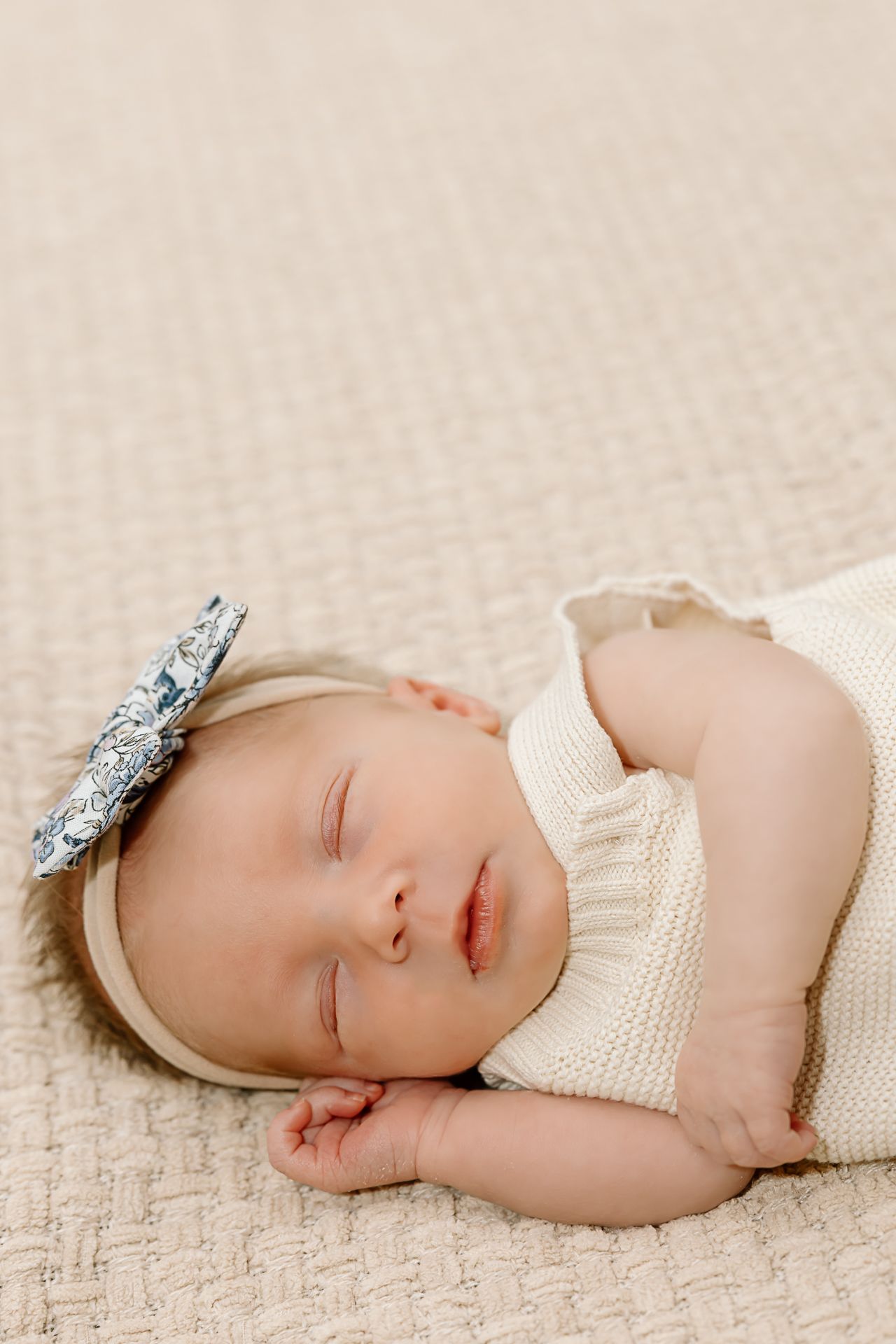 Peaceful sleeping newborn in cream knit outfit with sparkly bow headband on textured backdrop.