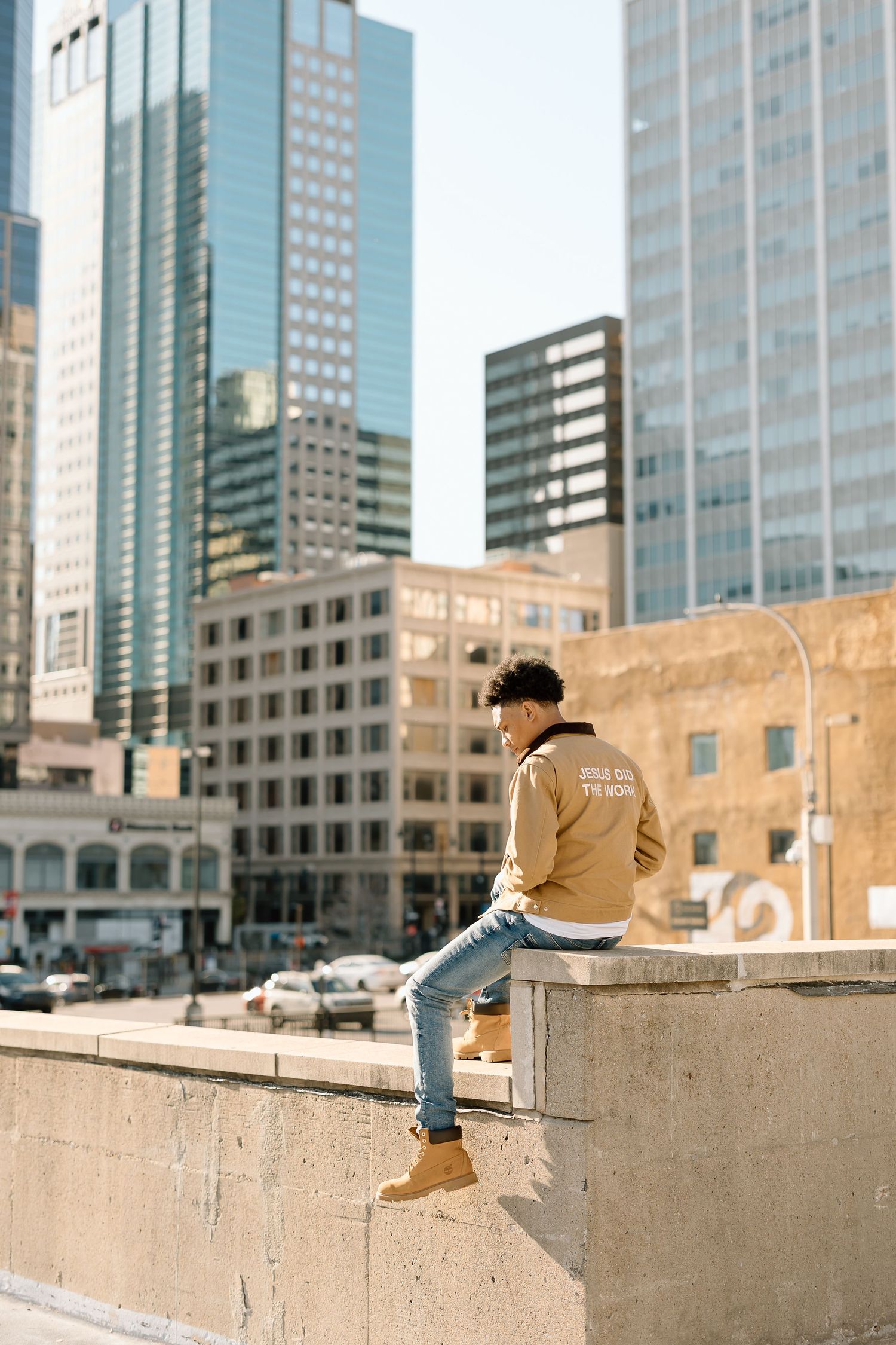 Person sits on concrete ledge overlooking downtown buildings on sunny day.