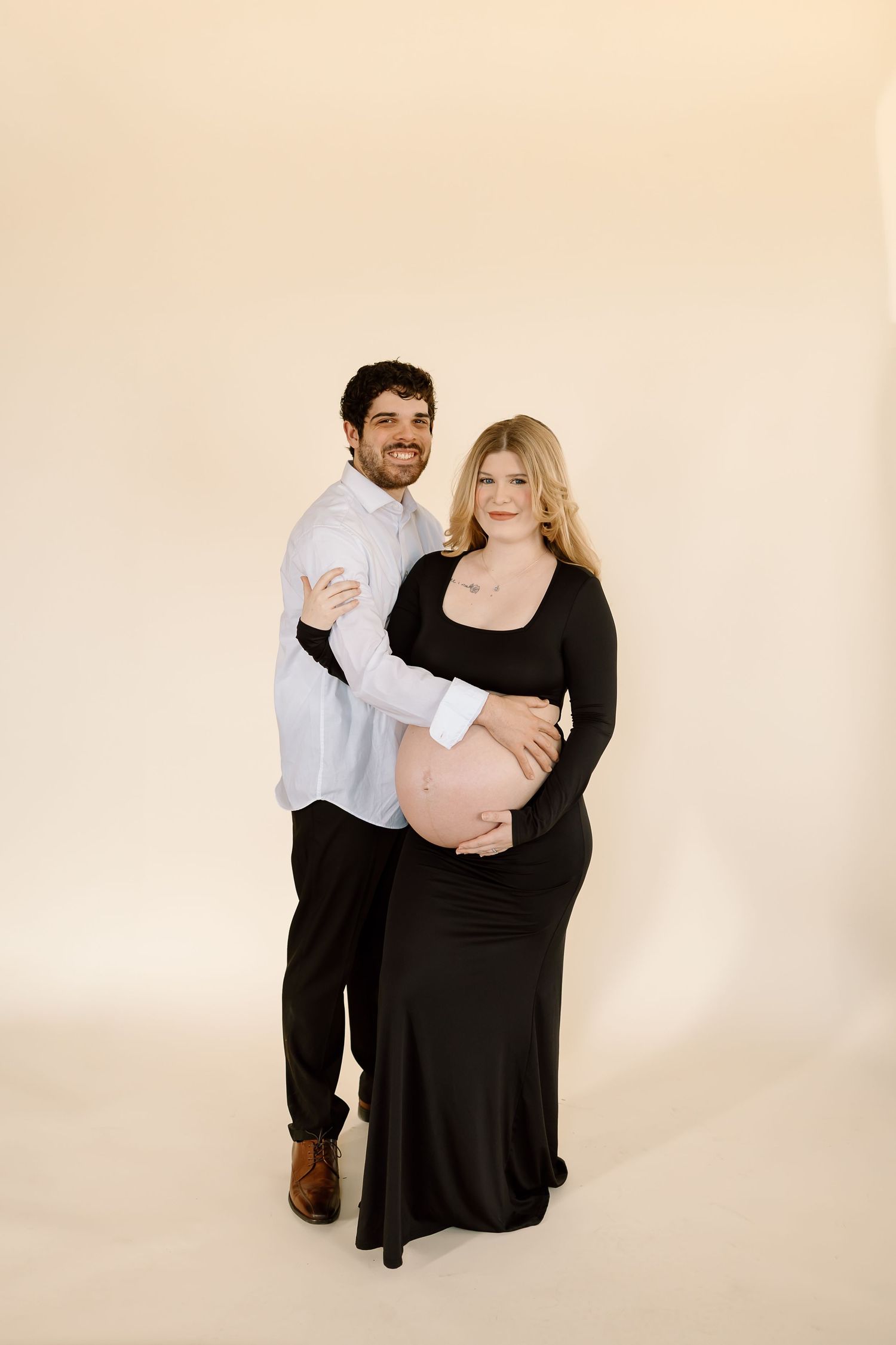 Sweet maternity studio portrait of couple in contrasting outfits against beige background.